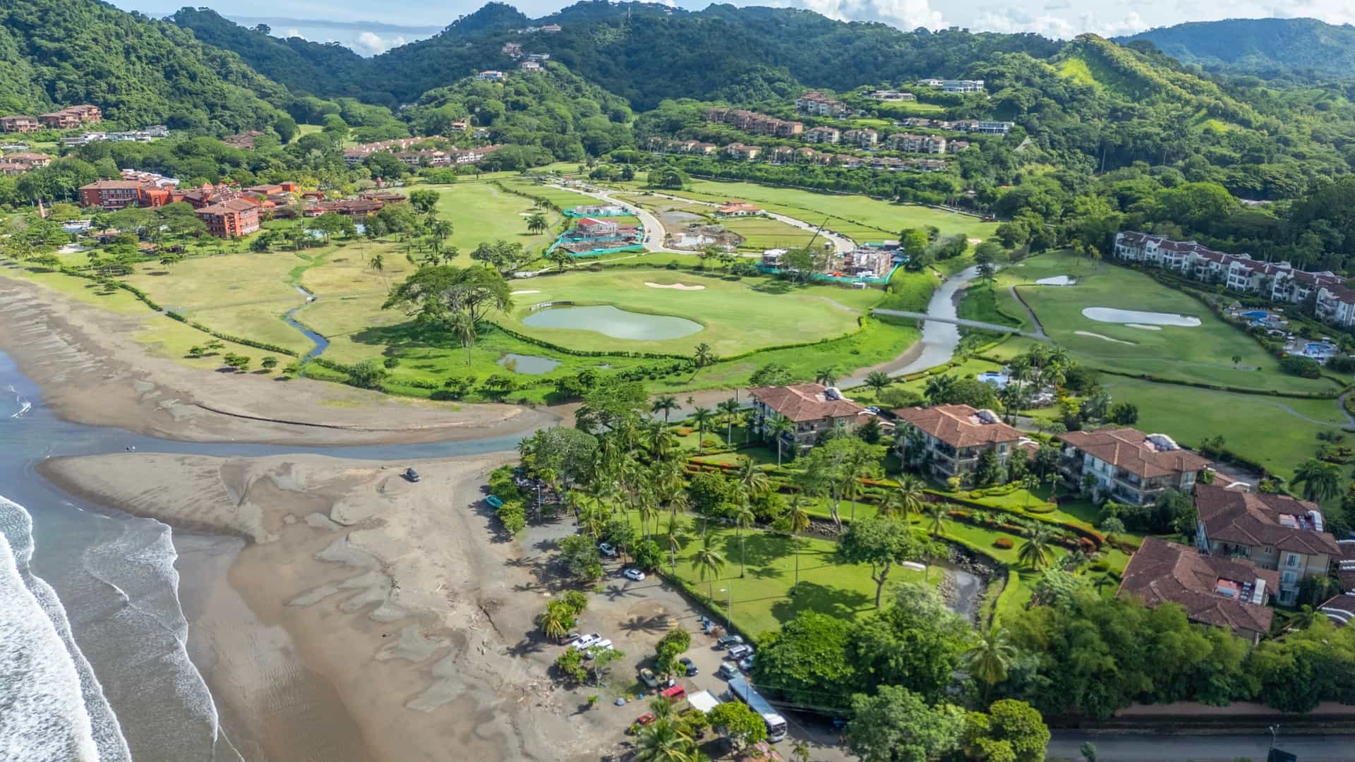 Aerial view of a resort area in Herradura, Costa Rica, featuring a lush green golf course, luxury residences with terracotta roofs, and a wide brown-sand beach bordering the Pacific Ocean.