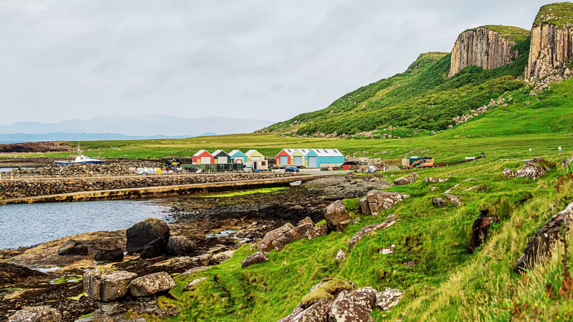 A breathtaking panoramic view of a dramatic, rugged landscape on the Isle of Skye, Scotland. The scene most likely depicts famous geological features such as the Quiraing or the Old Man of Storr on the Trotternish peninsula. The foreground is composed of lush green hills and a winding path, leading up to craggy, imposing rock formations and cliffs, all set against a vast sky, potentially with dramatic clouds or the soft light of sunrise/sunset.