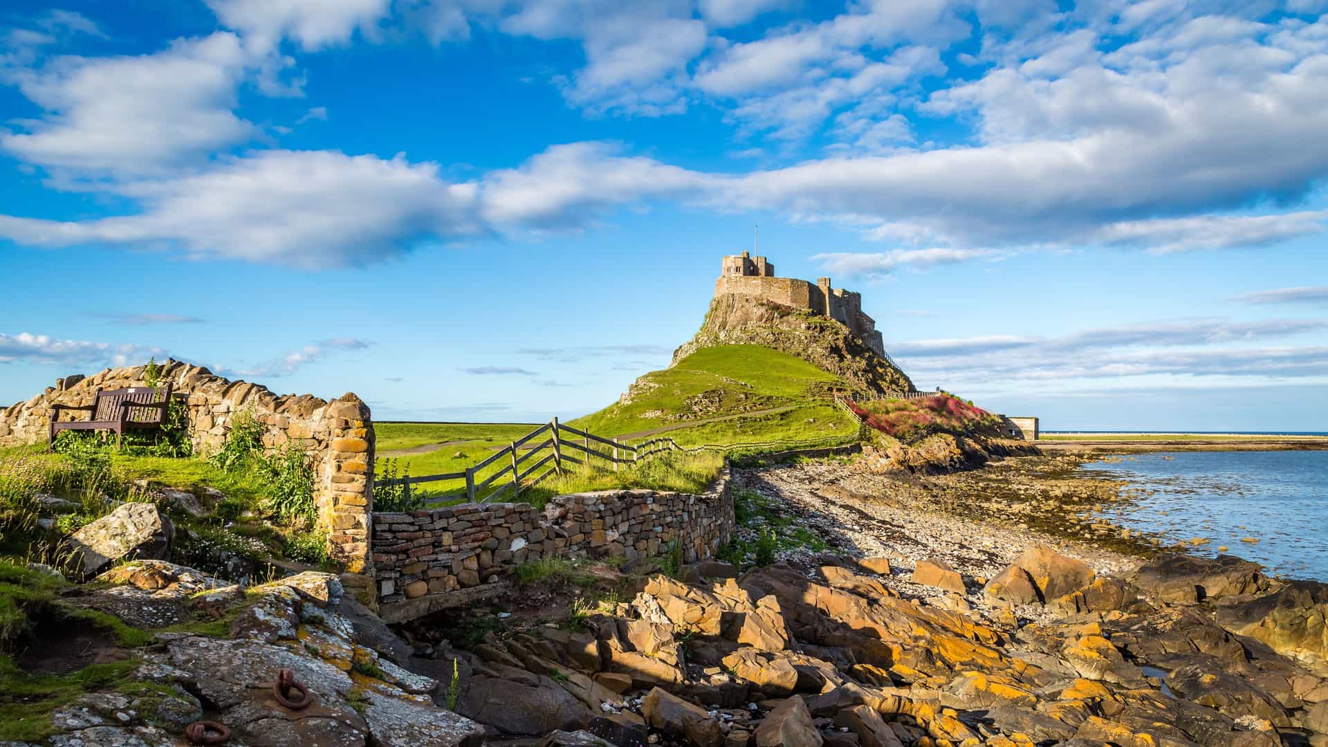 A stunning view of Lindisfarne Castle perched on a rocky outcrop, overlooking the sea on Holy Island, with a stone wall and bench in the foreground.