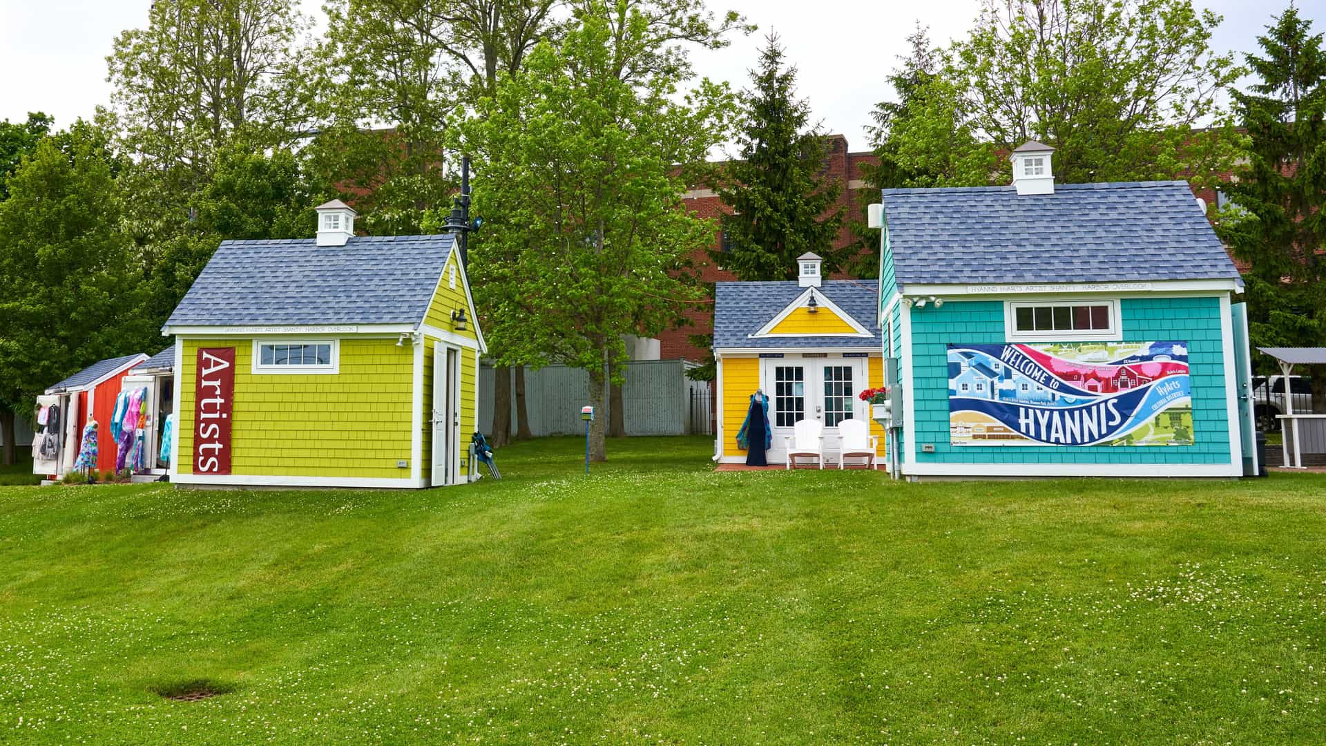 "A bright and cheerful row of colorful artist shacks on a green lawn with a large ""Welcome to Hyannis"" sign on one of the buildings in Hyannis, Massachusetts.  "