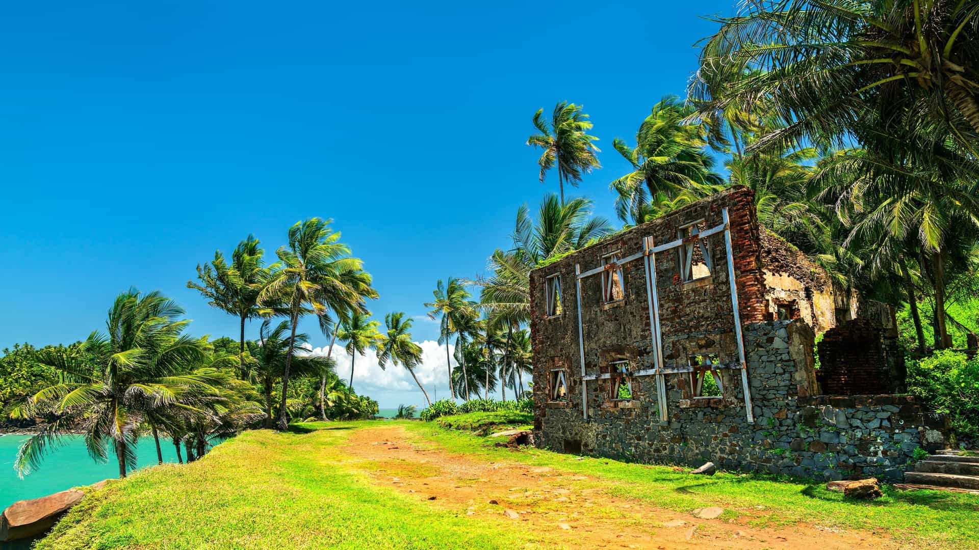 A vibrant tropical scene on Iles du Salut, French Guiana, with a large, ruined brick building next to a dirt path and surrounded by lush green grass, palm trees, and the turquoise ocean.