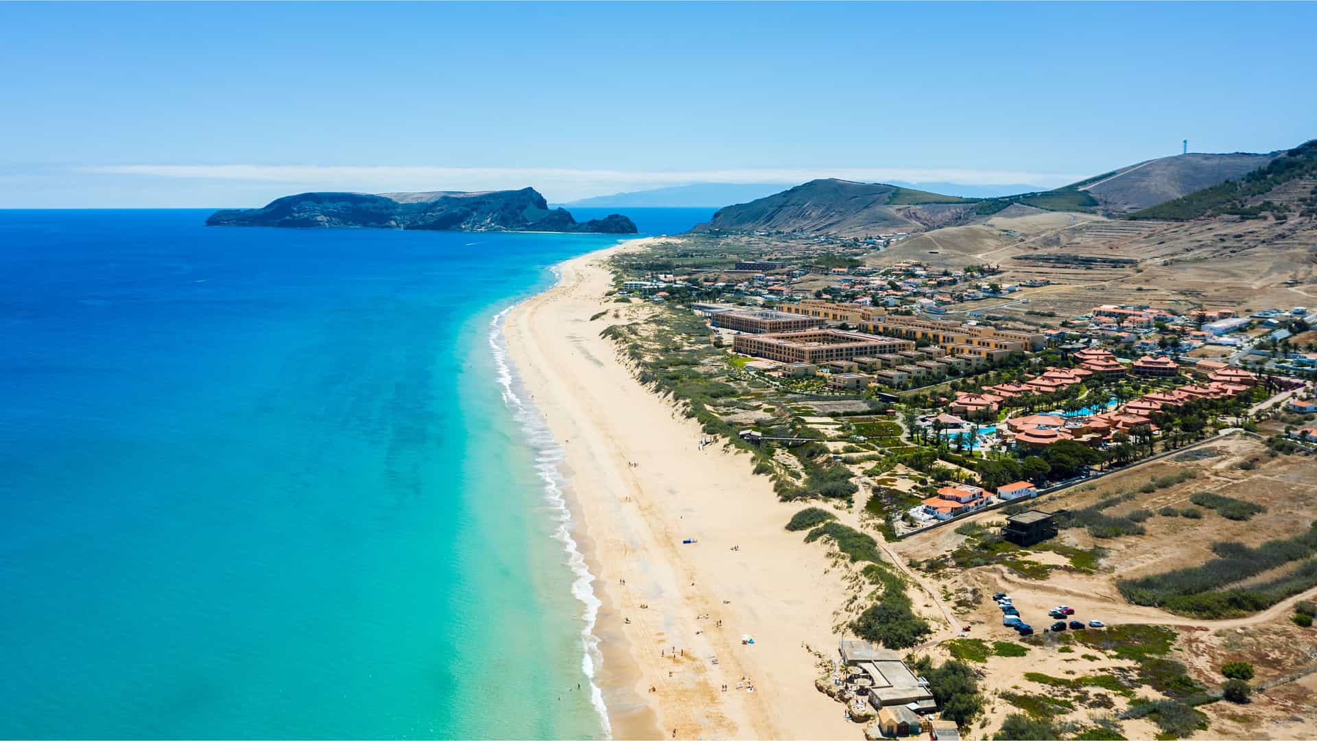 "An aerial view of Porto Santo Island, Portugal, showing the long, sandy beach and the turquoise waters of the Atlantic Ocean, with resorts and buildings nestled at the base of the island's mountains.  "