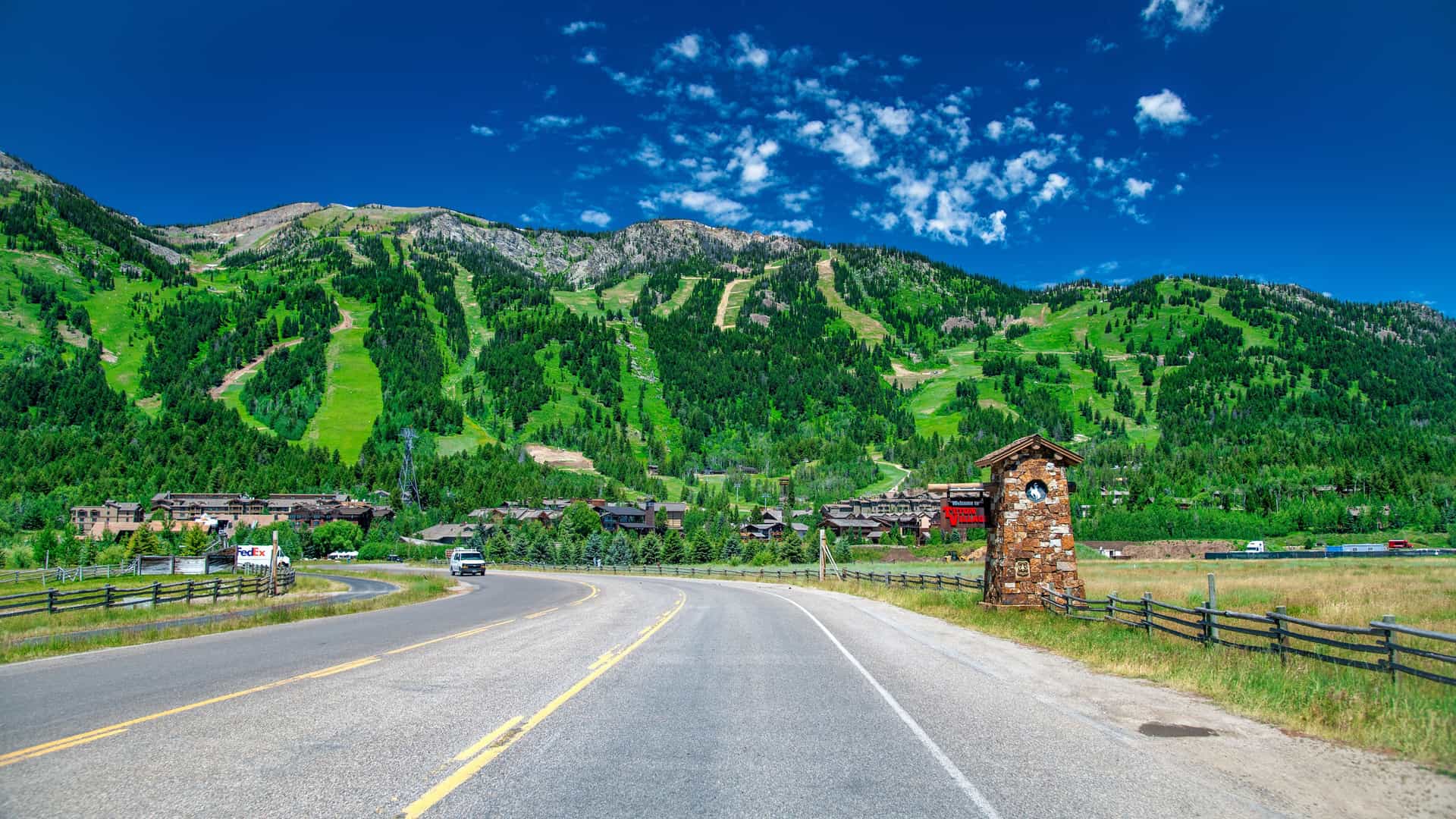 "A picturesque and winding two-lane road in Jackson Hole, Wyoming, leading towards the Teton mountain range with a prominent stone clock tower on the roadside.  "