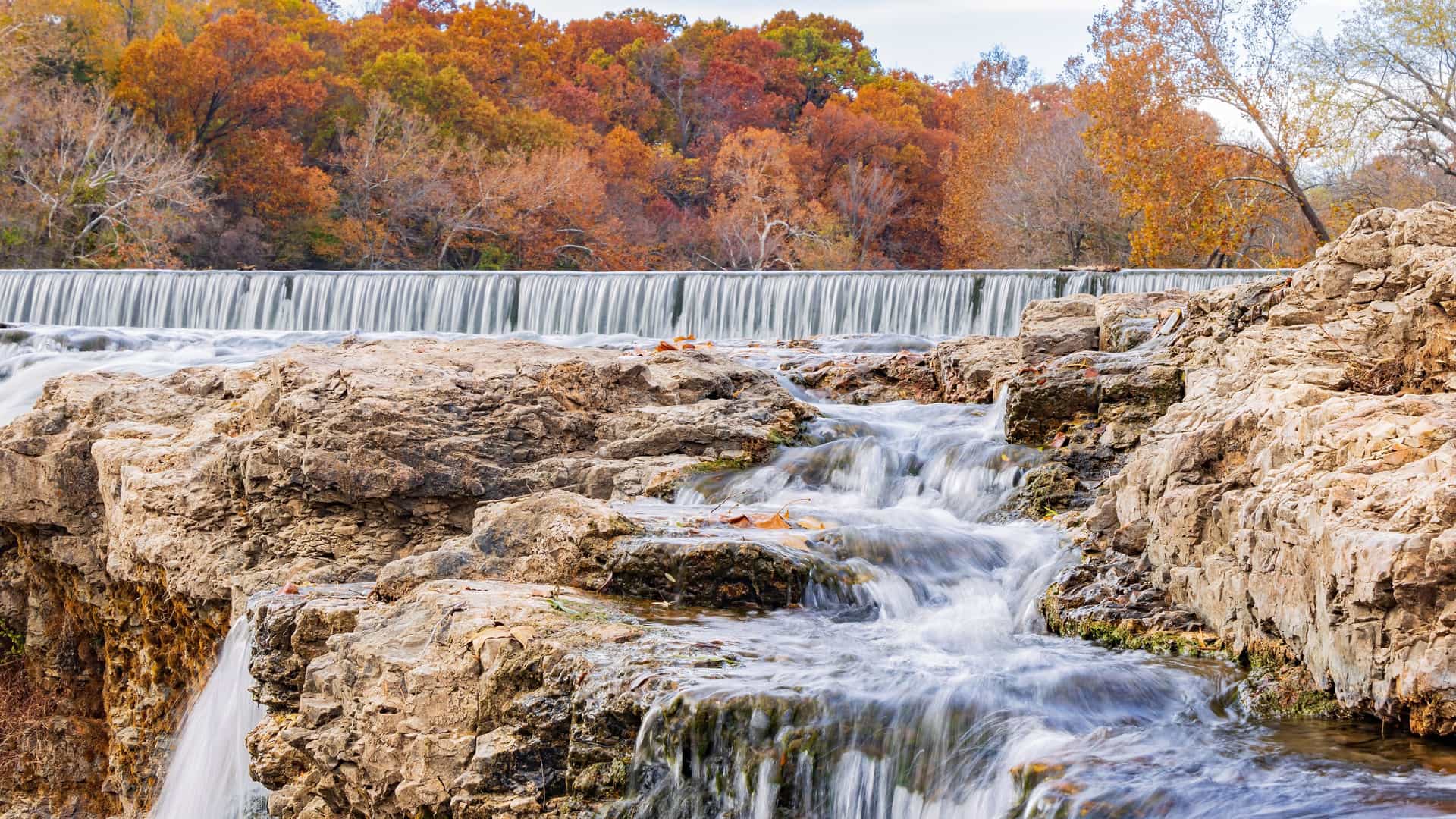 "A stunning autumn landscape shot of the Grand Falls waterfall on Shoal Creek in Joplin, Missouri, with water cascading over the rocky chert limestone formations and vibrant fall foliage in the background.  "