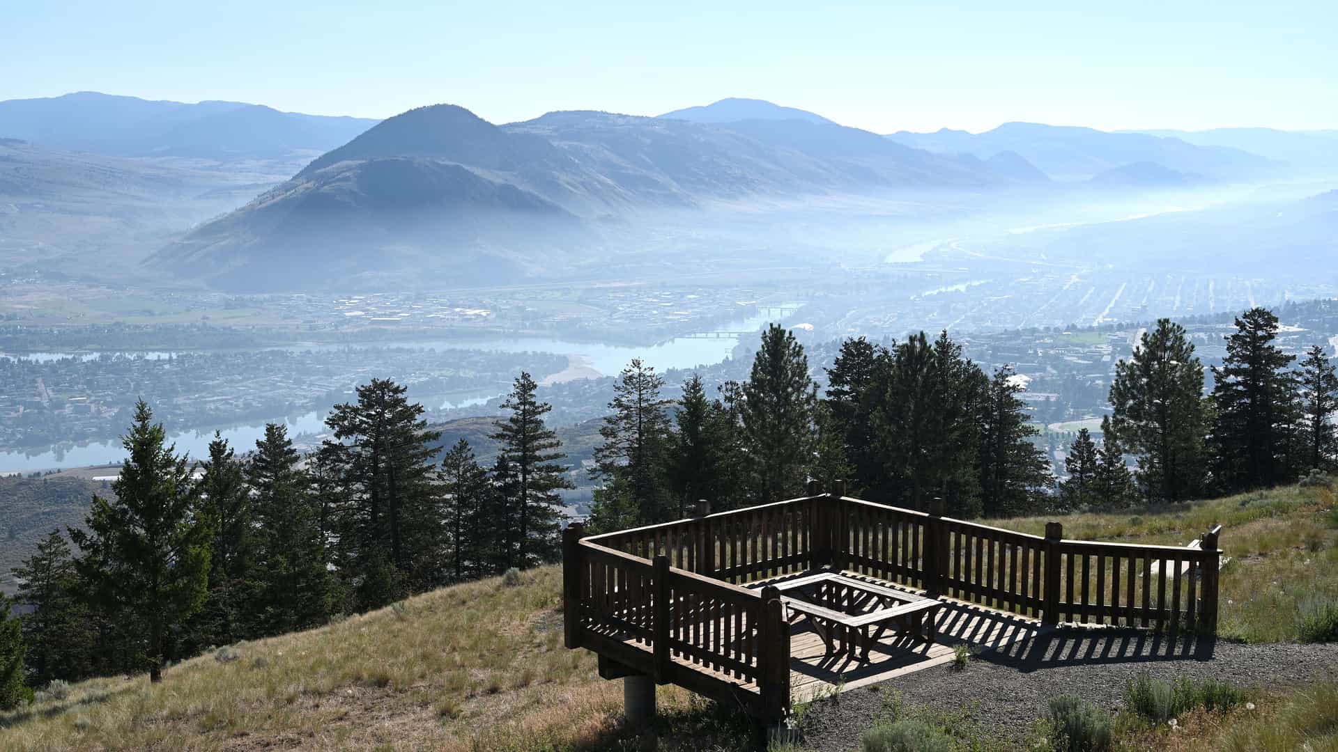 "A beautiful elevated panoramic view of the misty valley of Kamloops, British Columbia, with the North and South Thompson rivers flowing through the city, and a wooden viewing platform in the foreground.  "