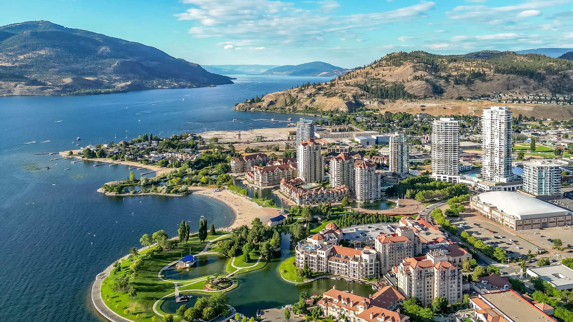"An aerial shot of Kelowna, British Columbia, showing the city's waterfront and marina on the shores of Okanagan Lake, with modern high-rises and residential buildings against a backdrop of forested hills.  "