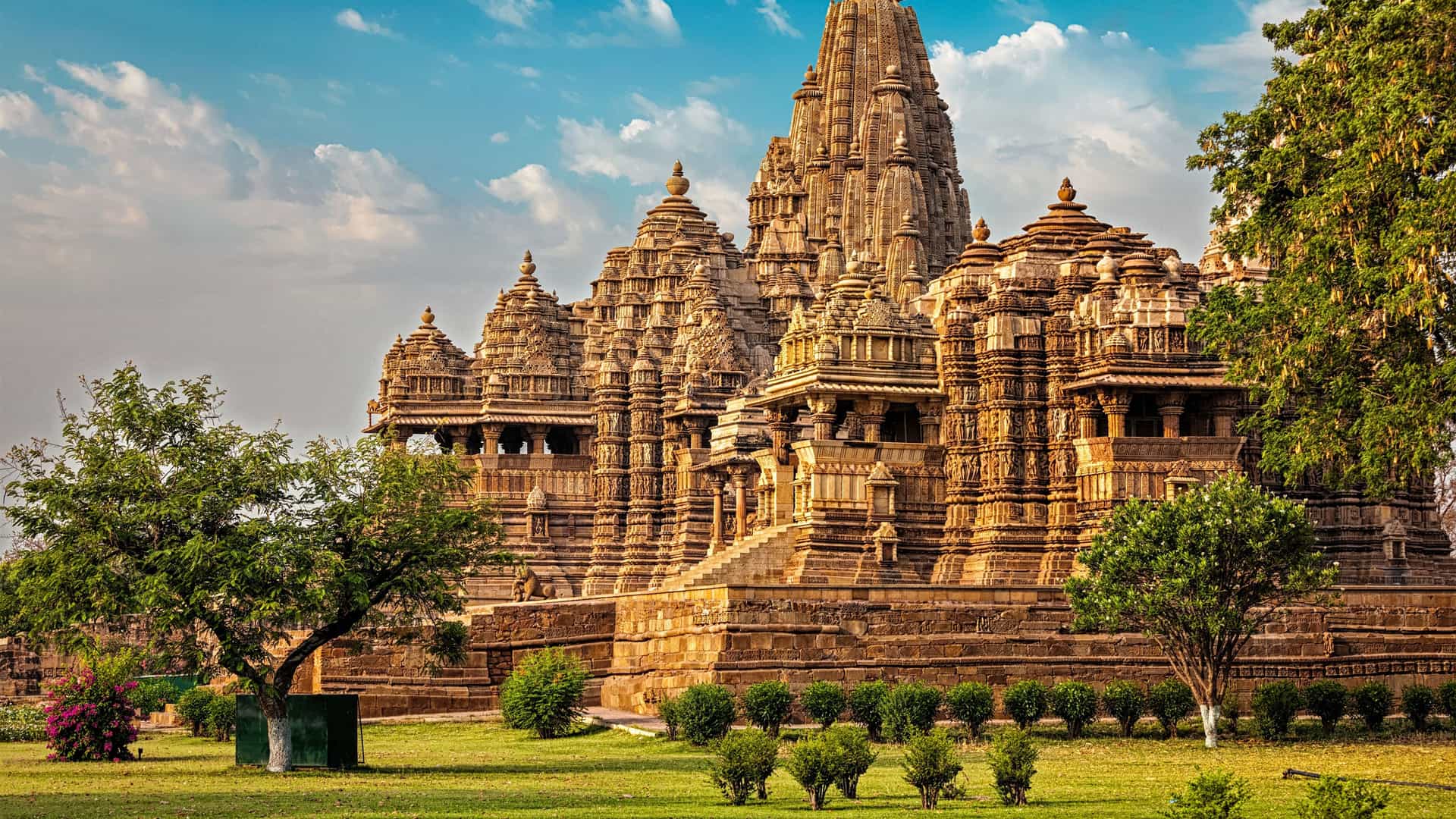 "A magnificent view of the Kandariya Mahadeva Temple, the largest of the ancient Hindu temples in Khajuraho, India, with its intricately carved spires and detailed stone architecture, surrounded by green lawns and trees.  "