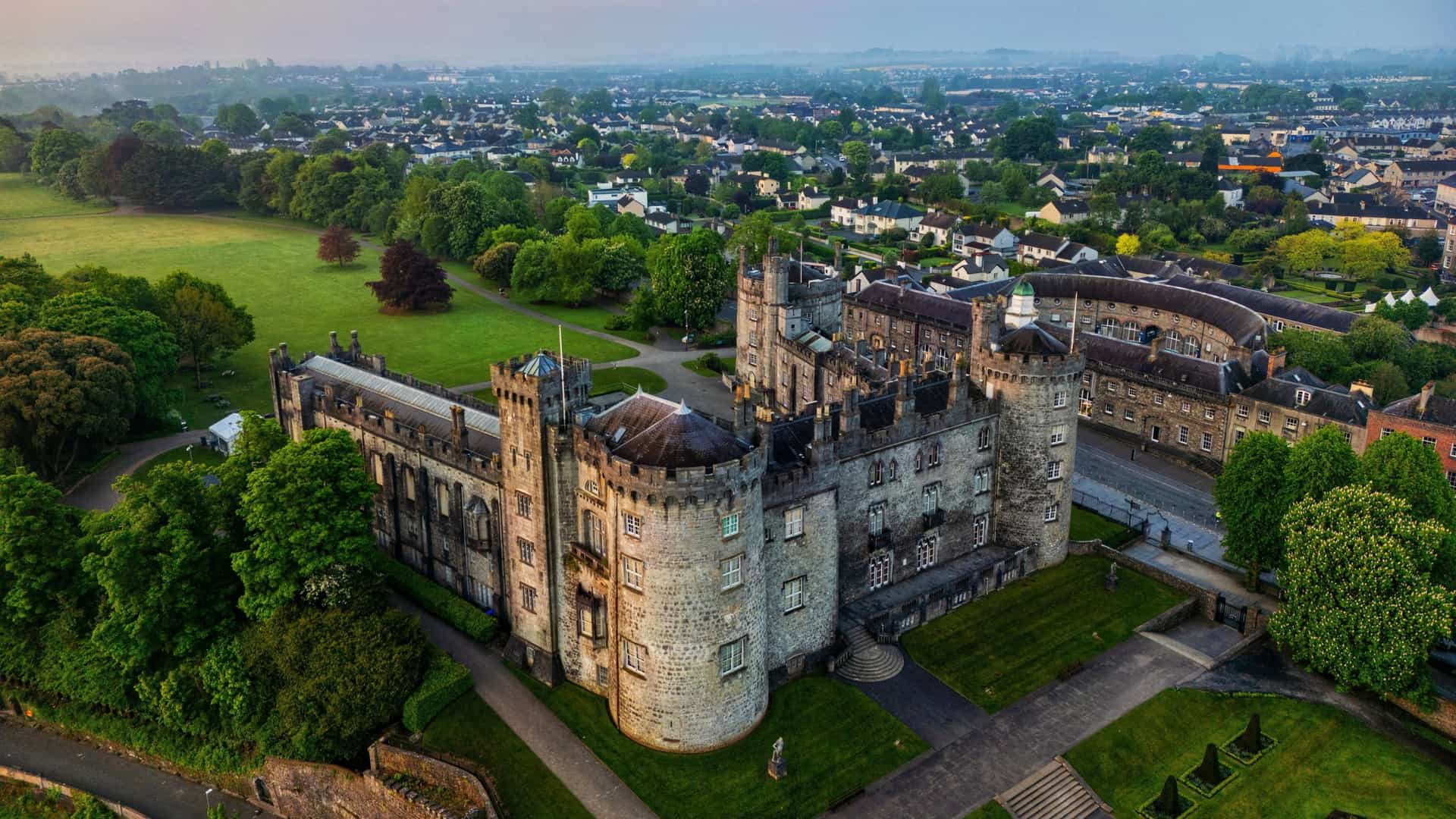 An aerial view of the historic Kilkenny Castle in Kilkenny, Ireland, featuring a large, medieval stone structure with rounded corner towers and manicured green lawns, set against the backdrop of the sprawling, low-rise town extending into the hazy distance.