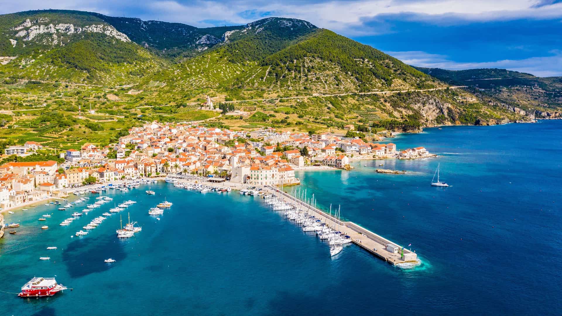 An aerial, panoramic view of the coastal town of Komiža on Vis Island, Croatia, in the Adriatic Sea. The picturesque town features tightly packed, historic stone buildings with orange and red tiled roofs, curving around a deep blue bay. Numerous sailboats, yachts, and fishing boats are moored in the harbor, and the town is dramatically backed by a large, steep, green mountain.