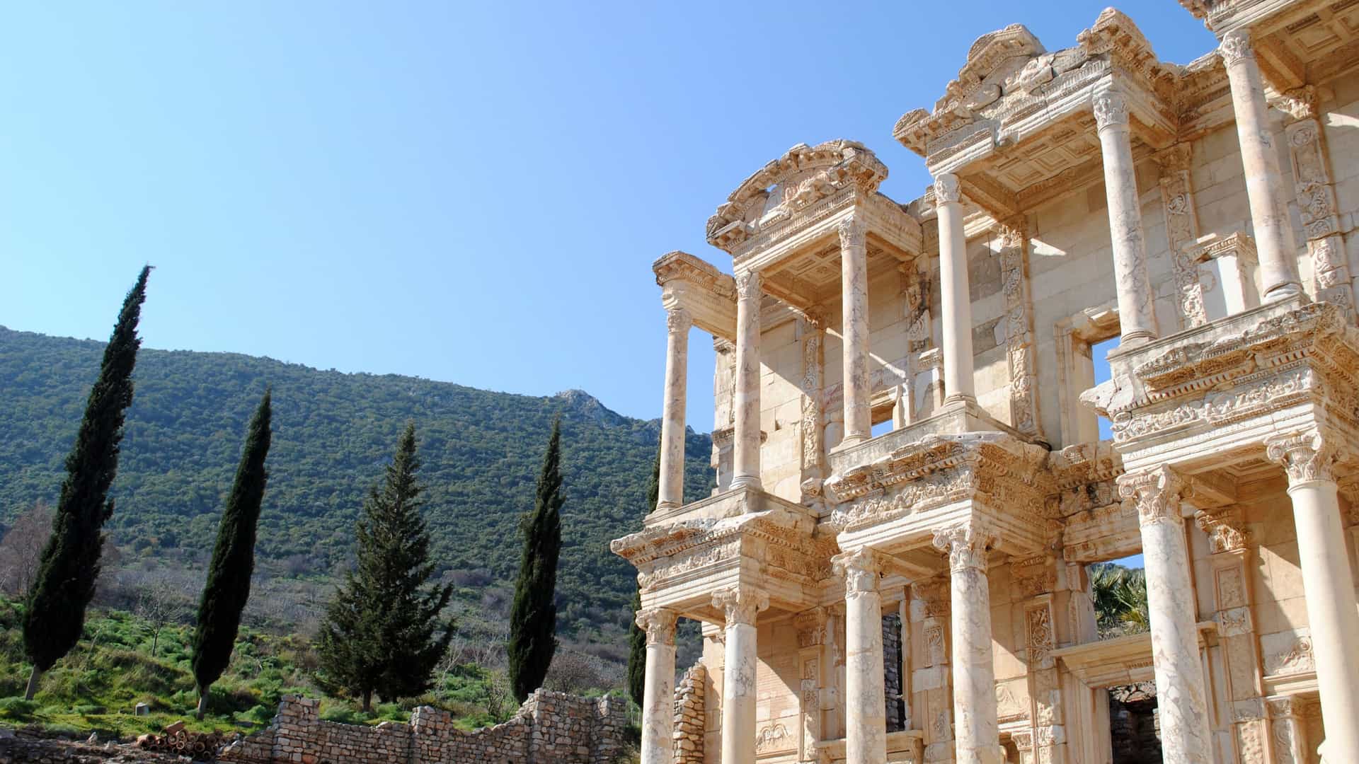 The iconic Library of Celsus in the ancient city of Ephesus, Turkey, with its magnificent, two-story facade and marble columns standing against a backdrop of cypress trees and green hills.