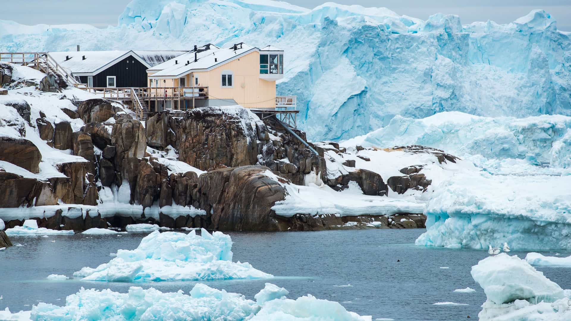 A picturesque scene in Kvanefjeld, Greenland, showing a yellow and a black house perched on a snow-covered rocky shore in front of a massive blue glacier.