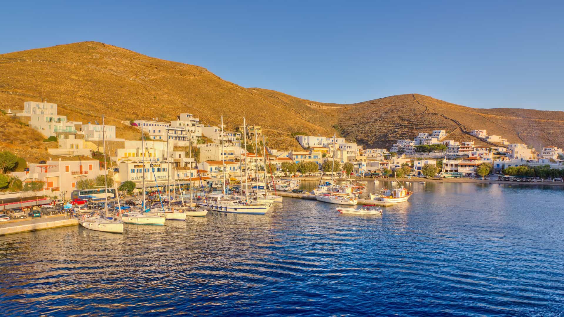 "A picturesque panoramic view of the Kythnos port in Greece, with white Cycladic houses and buildings lining the waterfront and a marina filled with sailboats, all nestled against the backdrop of dry, golden hills under a clear blue sky.  "