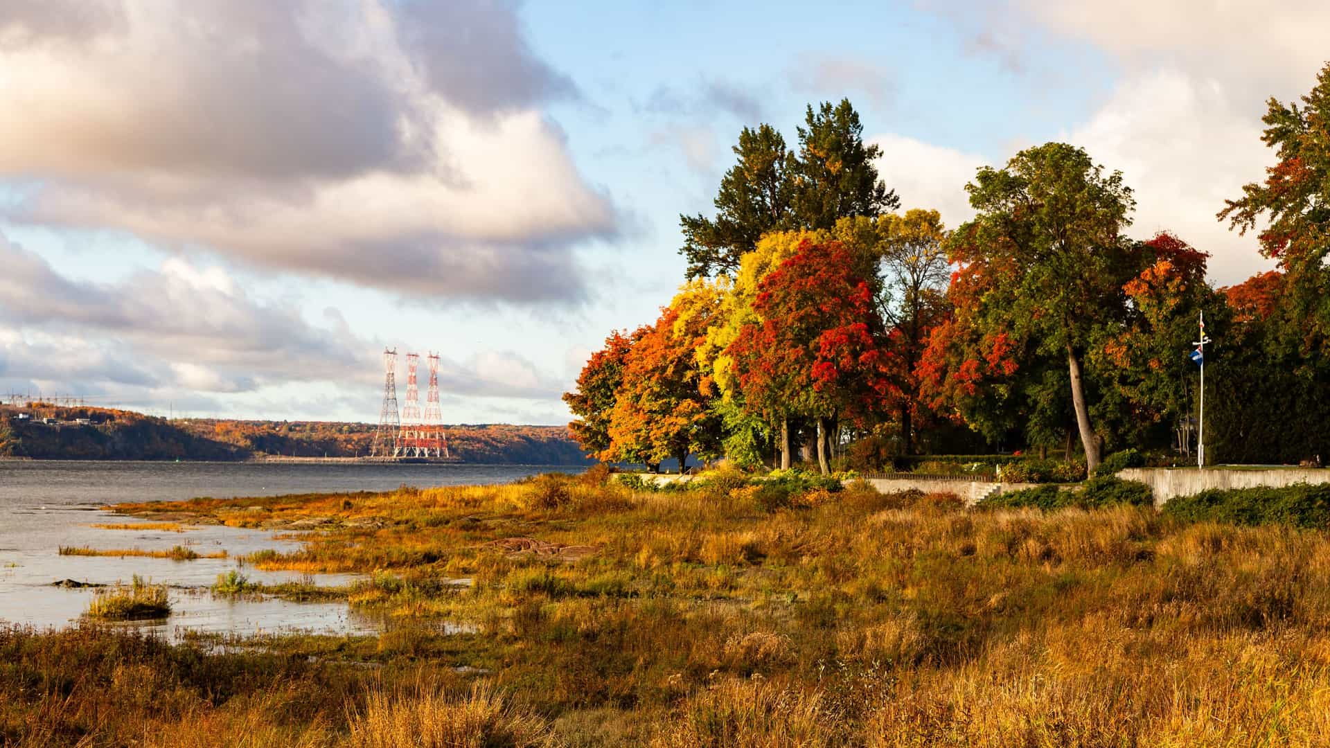 "A wide-angle landscape shot of the St. Lawrence River in L'Anse-au-Clair, Canada, showing a shoreline of marshy grass and a line of trees with vibrant autumn foliage.  "