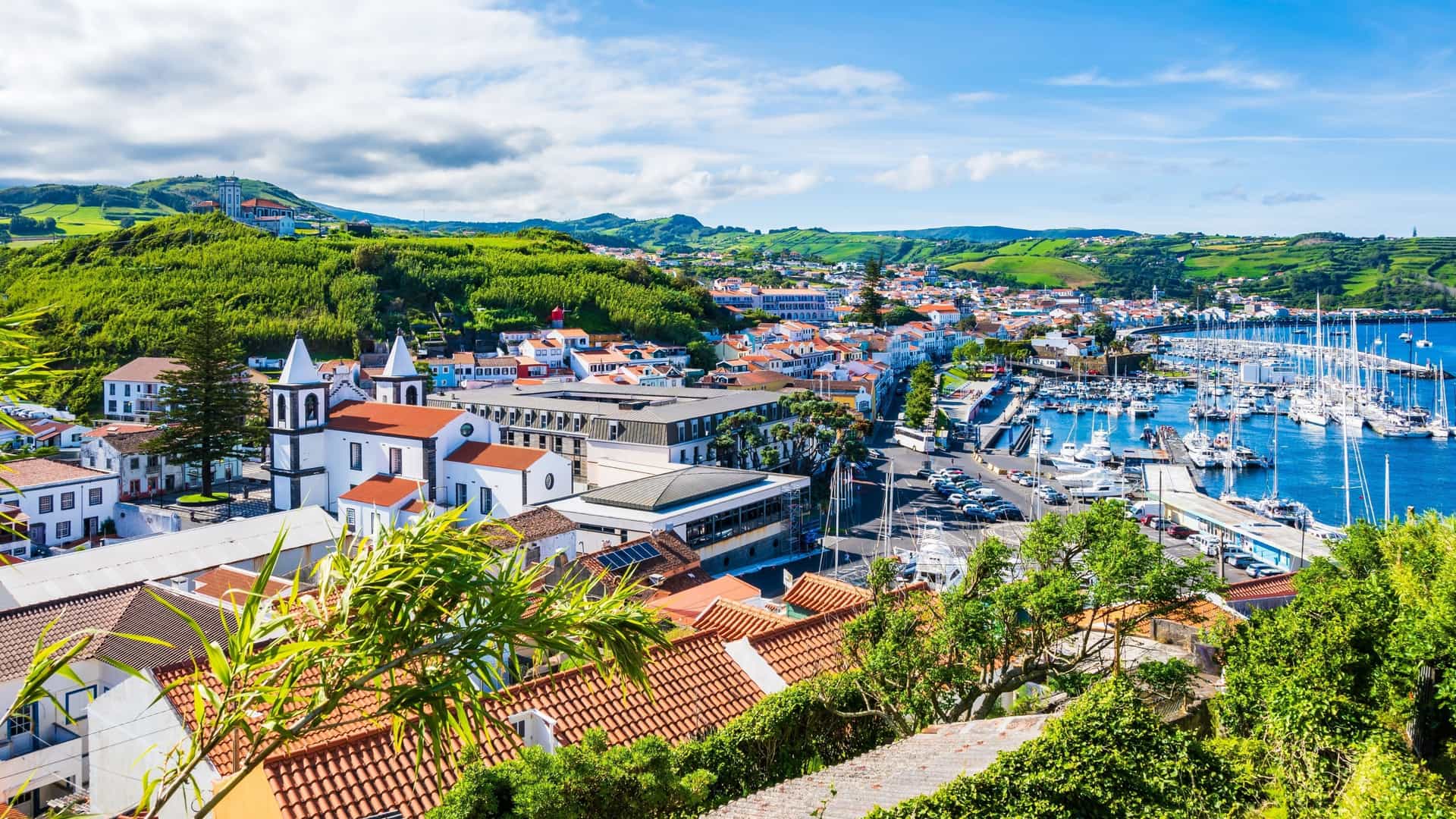 A scenic view of Horta, Azores, showing the harbor filled with sailboats, the white buildings of the city, and the lush green hills in the background.