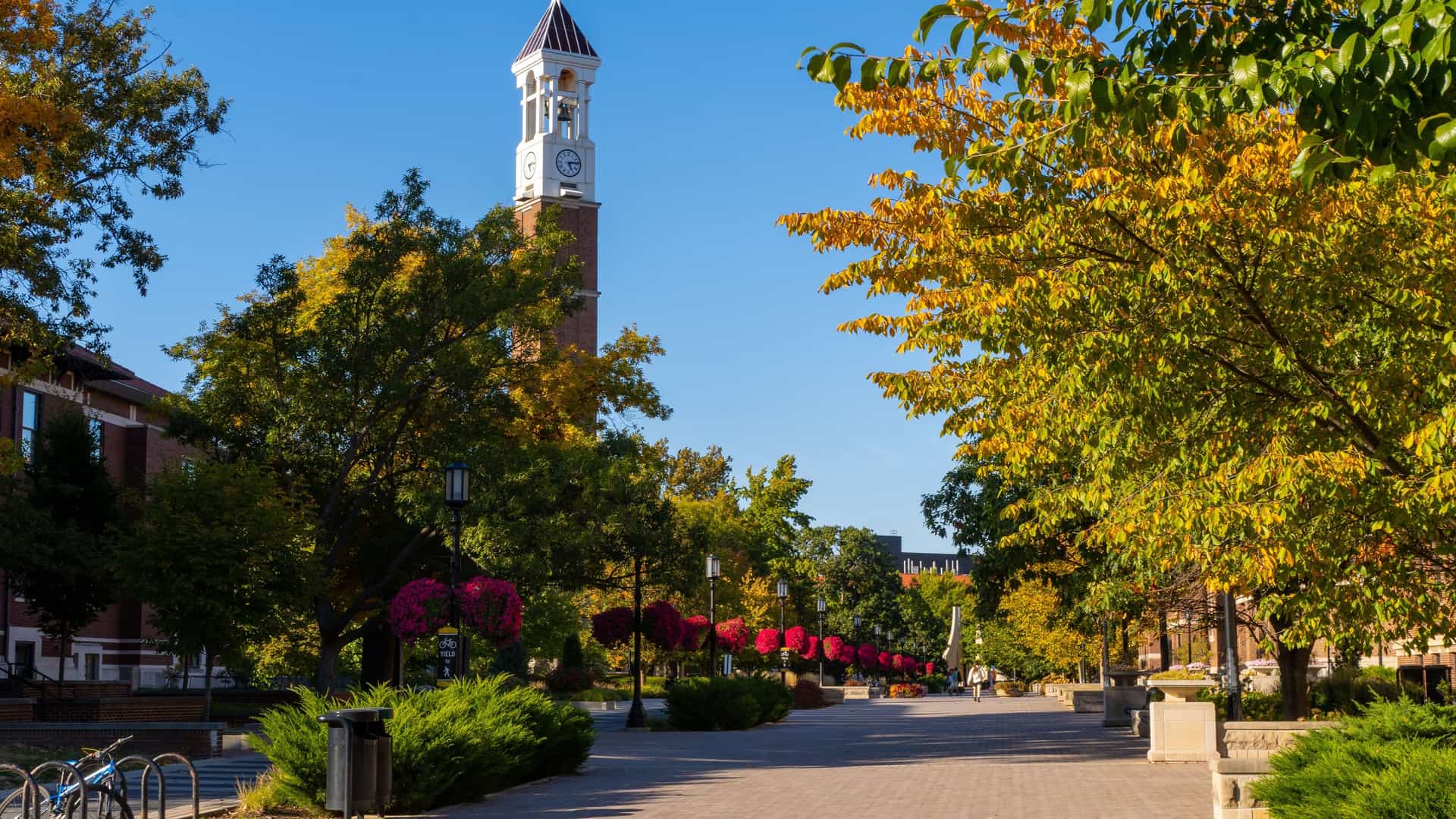 "A vibrant autumn scene on the campus of Purdue University in Lafayette, Indiana, showing the iconic Bell Tower and a path lined with trees in full fall foliage.  "