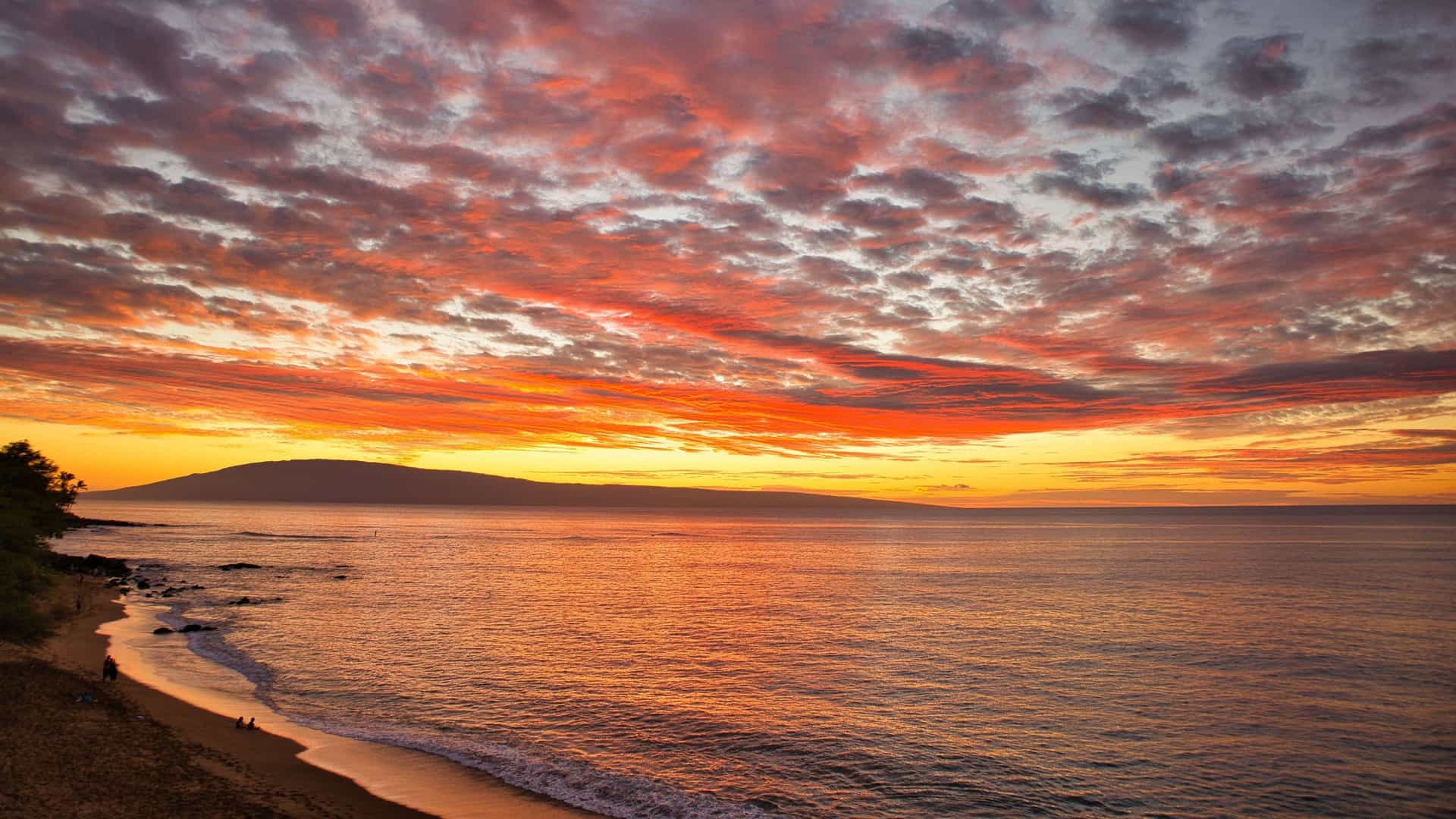 A vibrant sunset over the Pacific Ocean, with a fiery orange and red sky reflecting on the water and a dark silhouette of an island on the horizon near Lahaina, Maui.