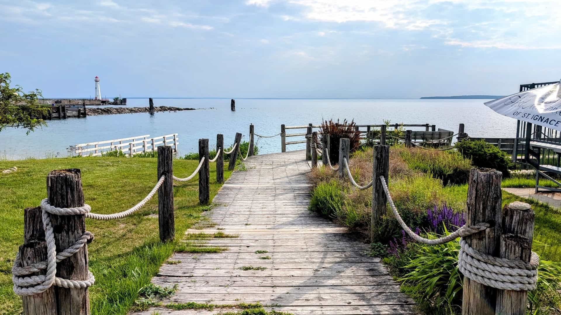 A wooden boardwalk path bordered by weathered wooden posts wrapped with thick rope leads toward the calm blue waters of Lake Huron on a partially cloudy day. In the distance to the left, a white lighthouse with a red cap is visible on a dock.