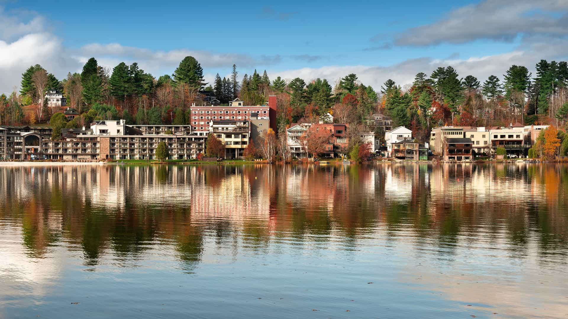 "A serene view of Mirror Lake in Lake Placid, New York, with hotels and houses on the shoreline reflecting in the still water, surrounded by vibrant fall foliage and green evergreen trees.  "