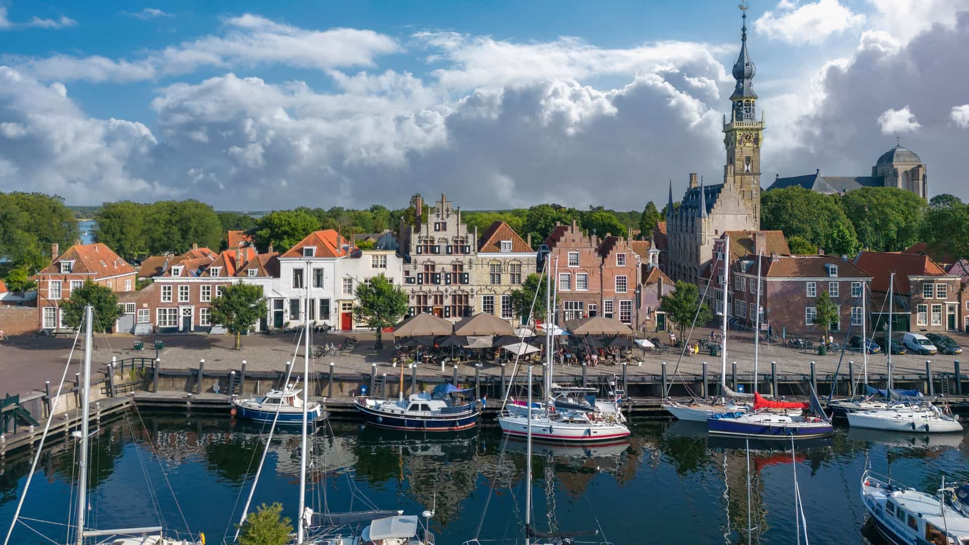 A serene waterfront scene in Lake Veere / Veerse Meer with calm marina waters reflecting docked sailboats, traditional gabled Dutch buildings, outdoor cafés, and a tall ornate clock tower framed by trees under a partly cloudy sky.