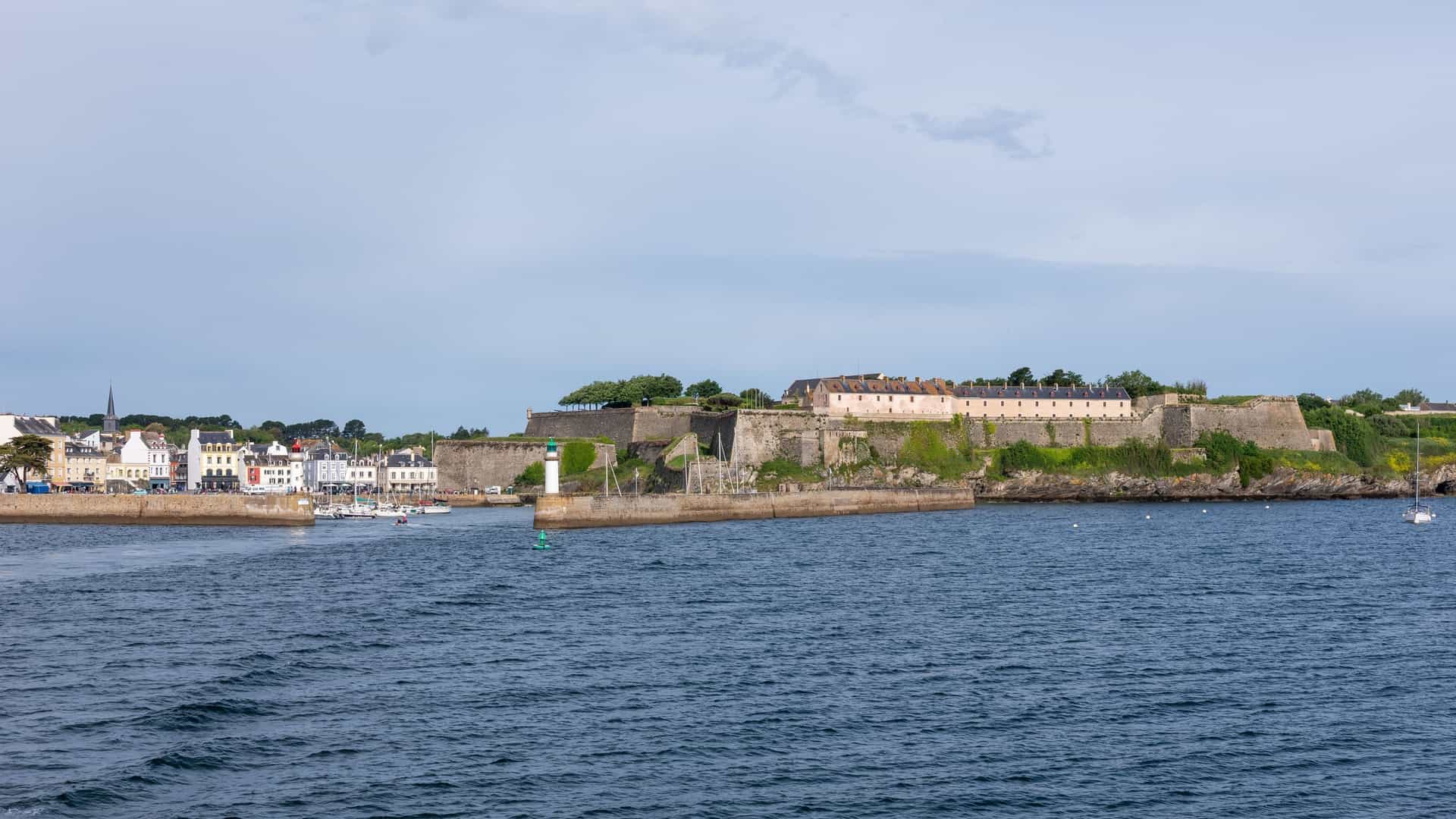 A scenic view of the harbor of Le Palais on Belle-Île-en-Mer, Brittany, France. The port is filled with moored boats in the foreground, with the colorful buildings of the town lining the waterfront. In the background, the imposing, star-shaped Vauban Citadel sits atop the headland, overlooking the harbor and the sea.