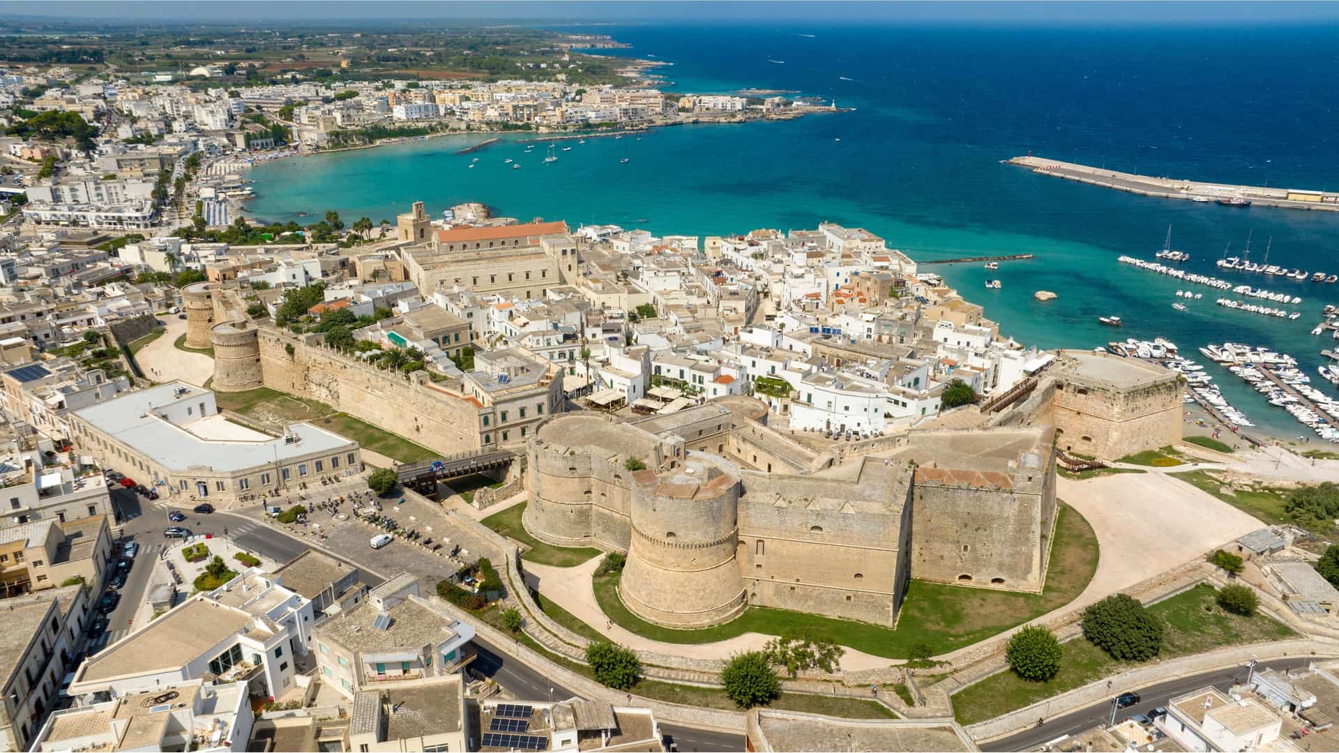 "An aerial view of the historic town of Otranto in the province of Lecce, Italy, showcasing the large coastal castle and the marina with boats in the turquoise sea.  "