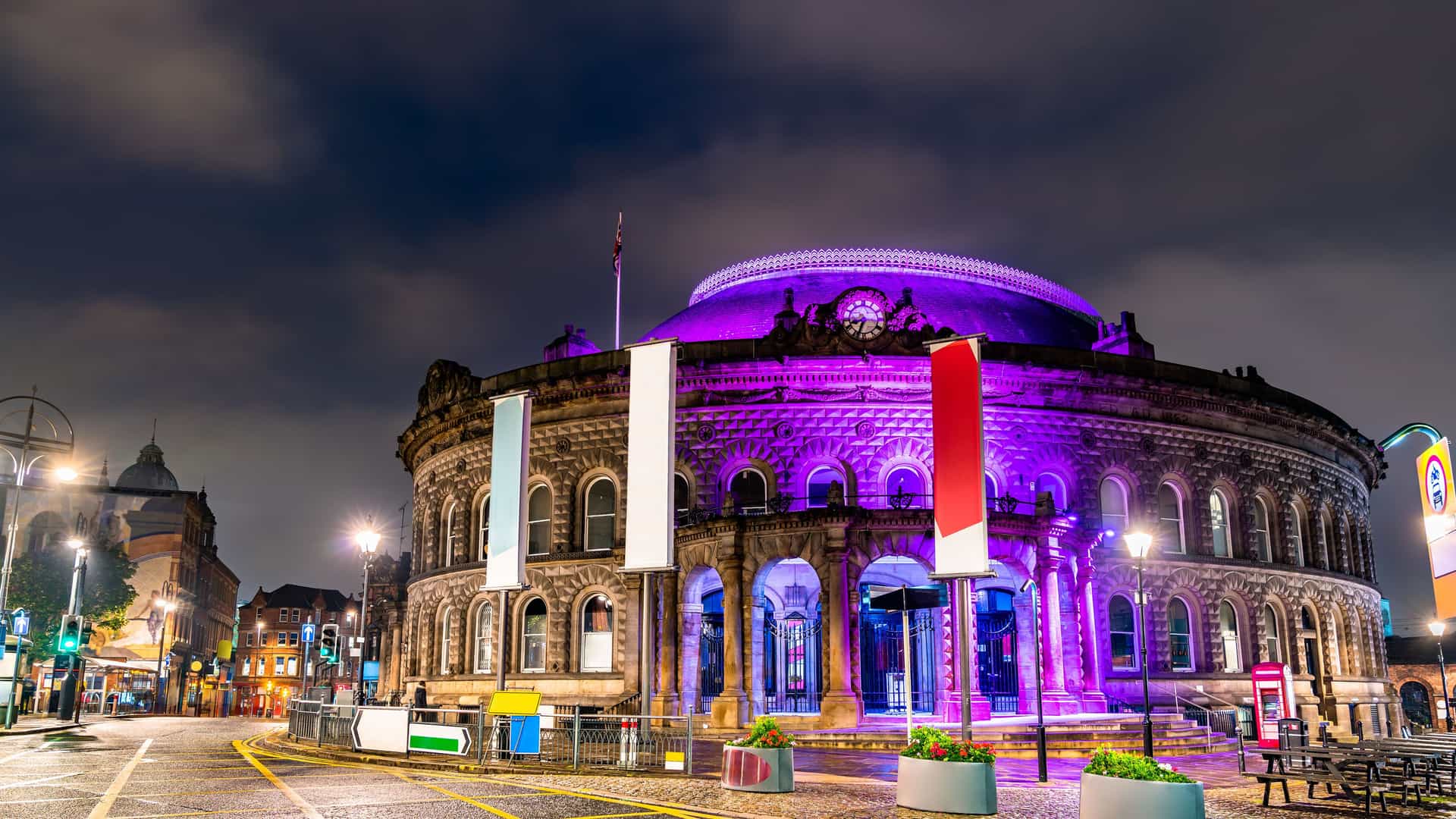 "A long exposure night shot of the Leeds Corn Exchange in Leeds, England, illuminated with vibrant purple lights under a cloudy sky.  "