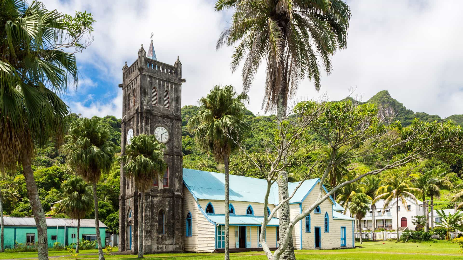 A scenic view of a historic church with a stone clock tower and a light blue roof, surrounded by lush tropical vegetation and palm trees, with a mountain range in the background on Leleuvia Island, Fiji.
