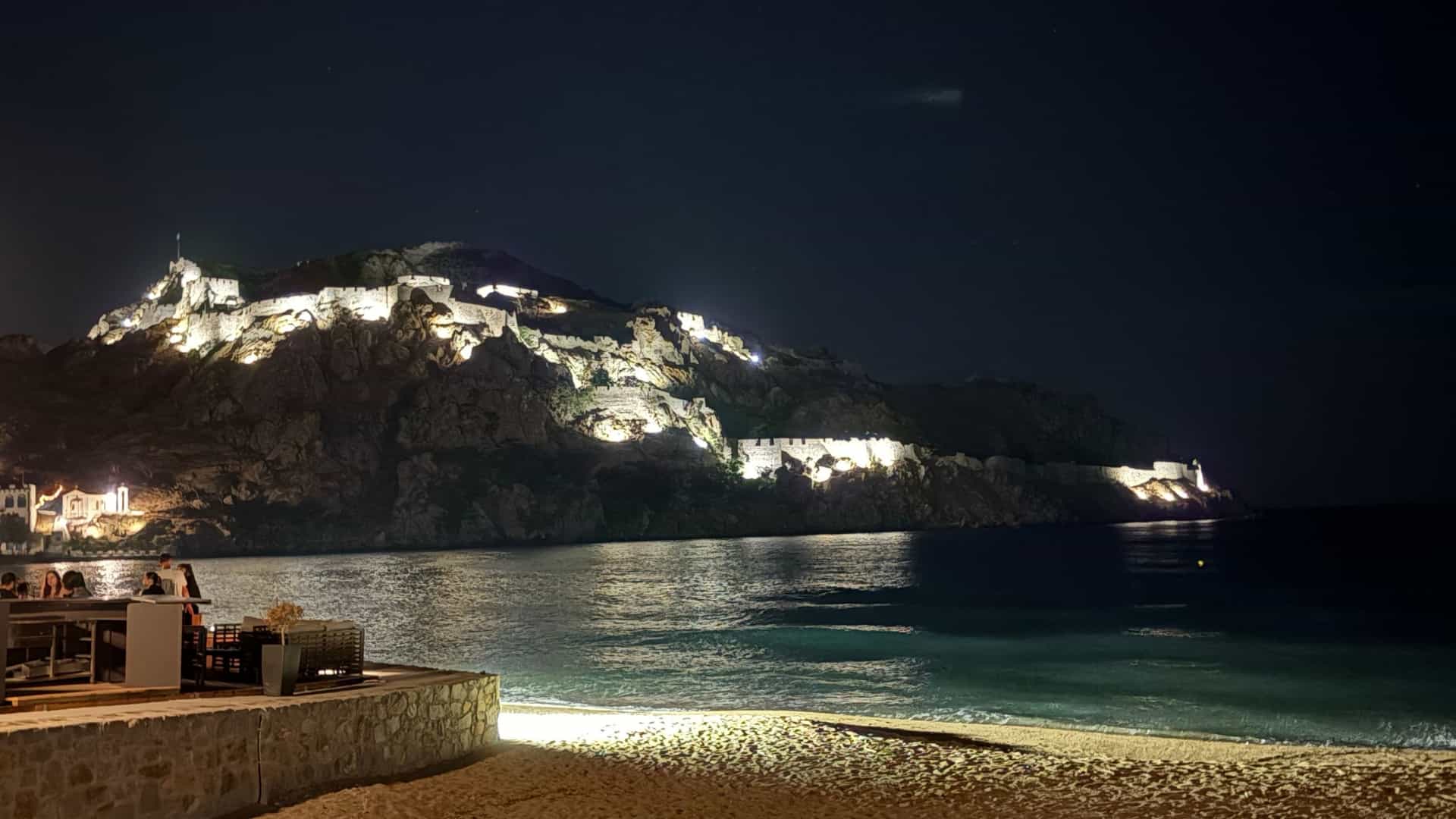 A dramatic nighttime view of the brightly lit Kastro (Castle) fortress walls on a steep, rocky headland overlooking the dark water and a sandy beach in Myrina, Lemnos, Greece. Several people are visible sitting at an outdoor restaurant on the left in the foreground.