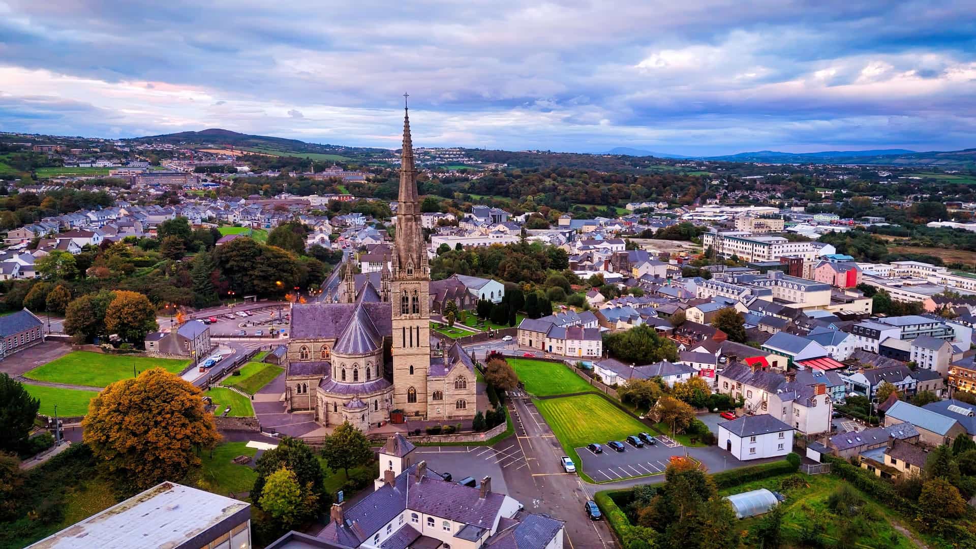 "An aerial view of Letterkenny, Ireland, showing the neo-gothic St. Eunan's Cathedral with its tall spire, surrounded by the town's buildings and green landscapes at sunset.  "