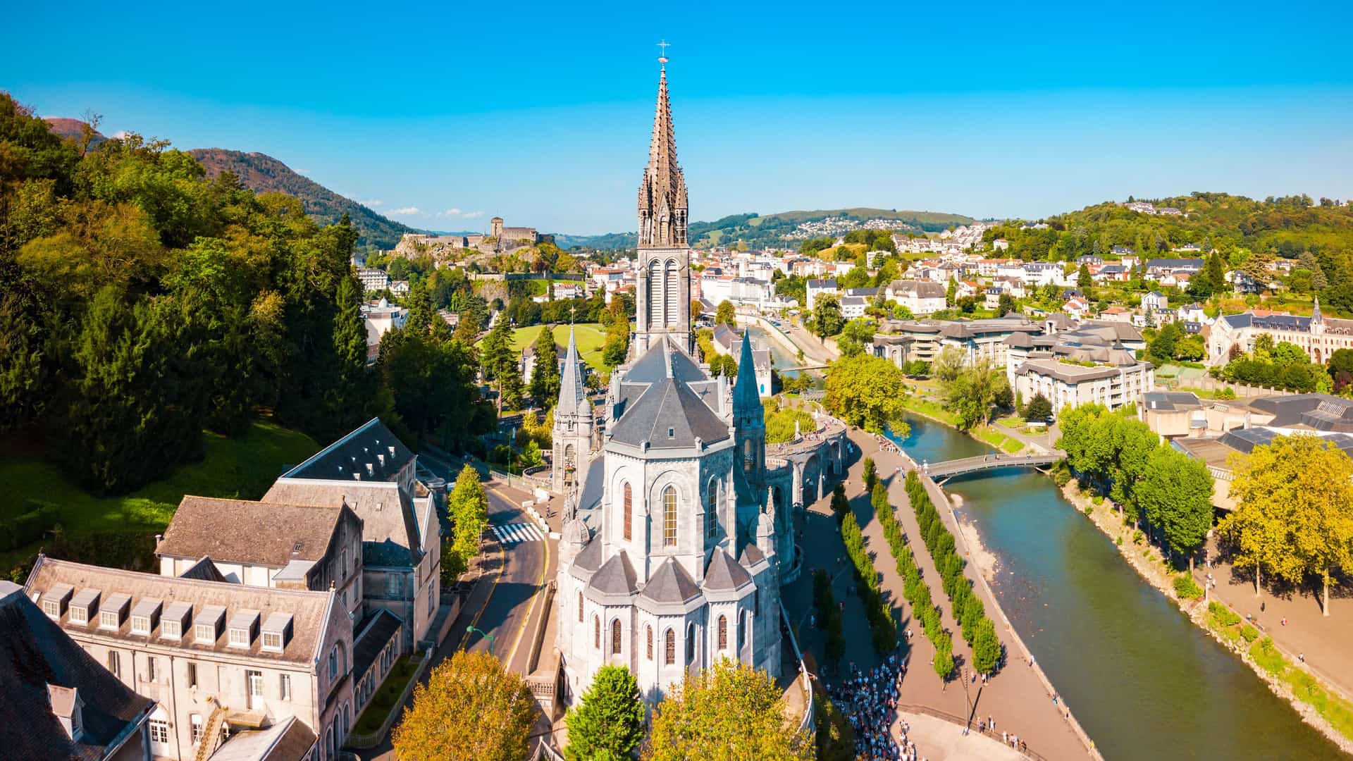 "An aerial panoramic view of the Sanctuary of Our Lady of Lourdes in Lourdes, France, showing the famous basilica, the Gave de Pau river, and the surrounding Pyrenees mountains.  "