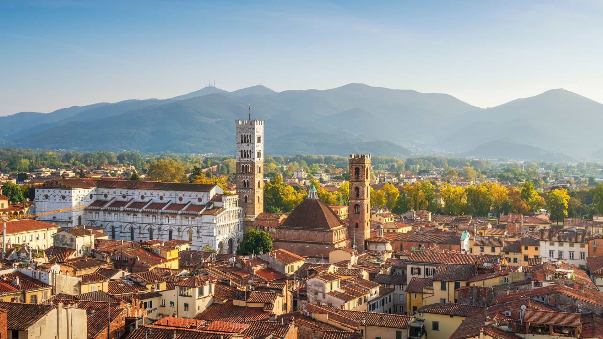 "An aerial view of the historic city of Lucca, Italy, with its terracotta rooftops, prominent bell towers, and a backdrop of rolling Tuscan hills.  "
