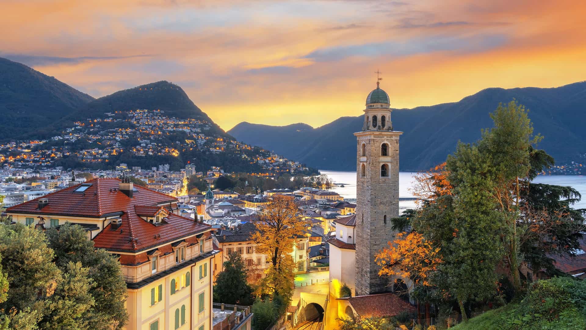 "An elevated sunset view of Lugano, Switzerland, highlighting the beautiful bell tower of the Church of Santa Maria degli Angeli, with Lake Lugano and the surrounding mountains in the background.  "
