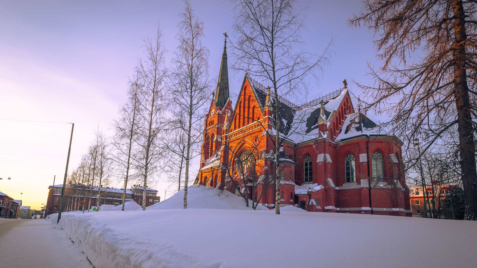 A winter image of the red-brick Luleå Cathedral in Sweden, covered in deep snow and surrounded by bare trees, glowing in the warm light of the setting sun.