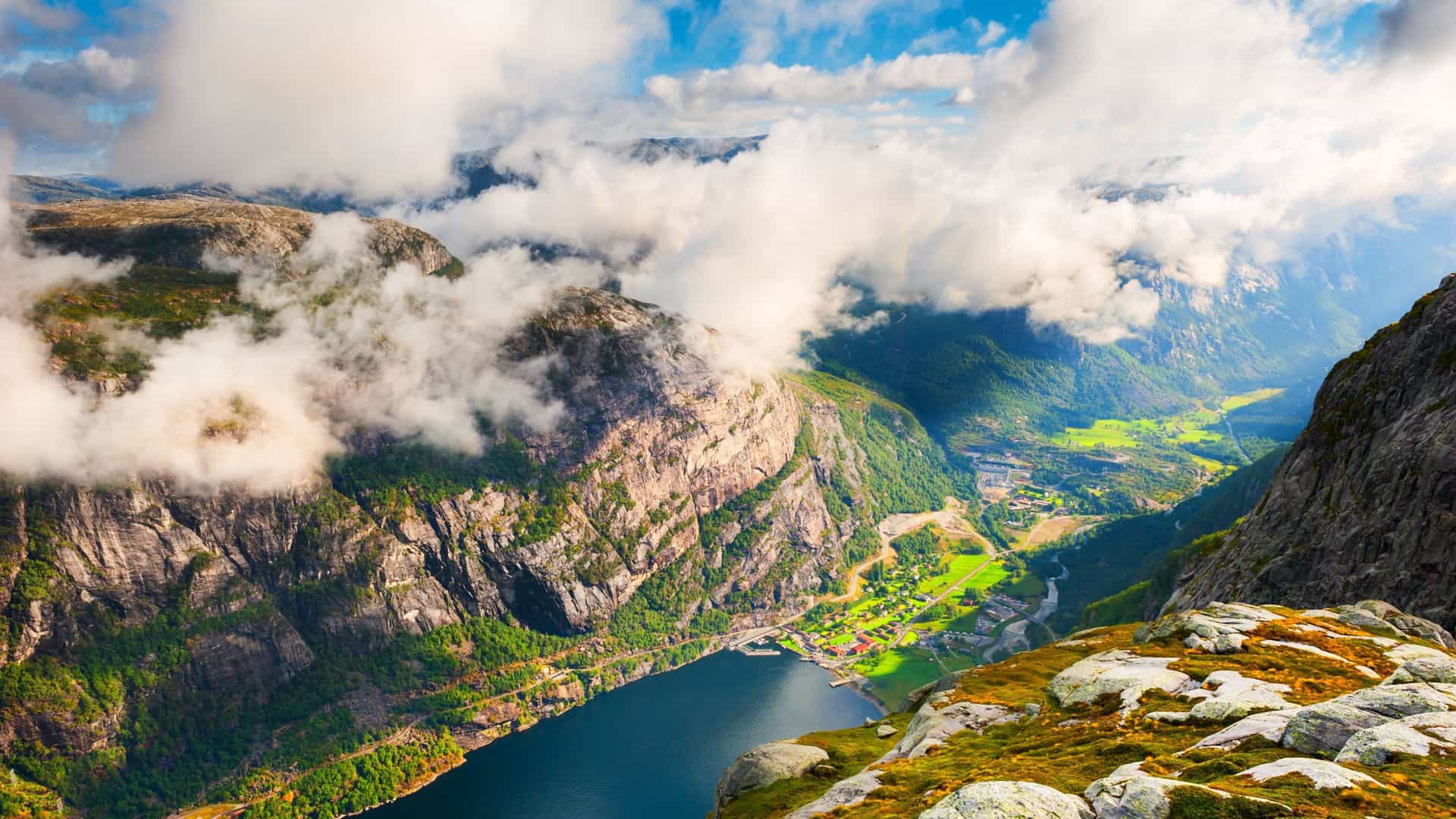 A high-angle panoramic view of the dramatic Lysefjord in Norway, with a small village nestled on the fjord's edge, surrounded by steep mountains and low-hanging clouds.