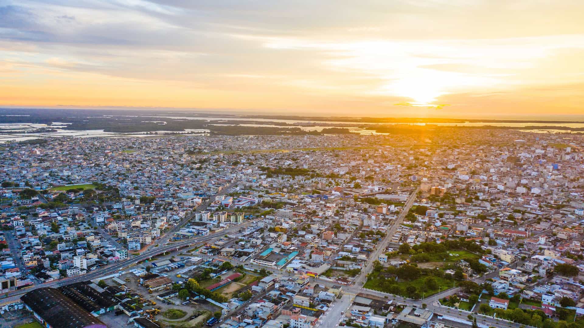 An aerial panoramic shot of the port city of Machala, Ecuador, showing the vast cityscape stretching toward the coast with a beautiful sunset in the background.