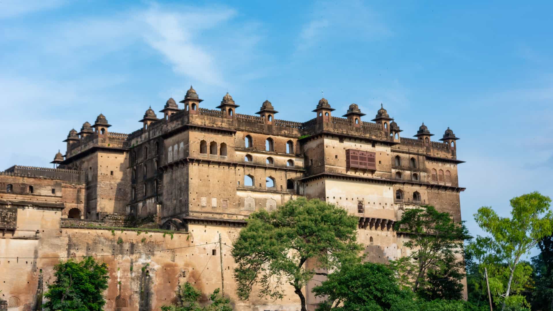 The historic Orchha Palace stands tall with intricate architecture and domes, set against a blue sky, surrounded by green trees.