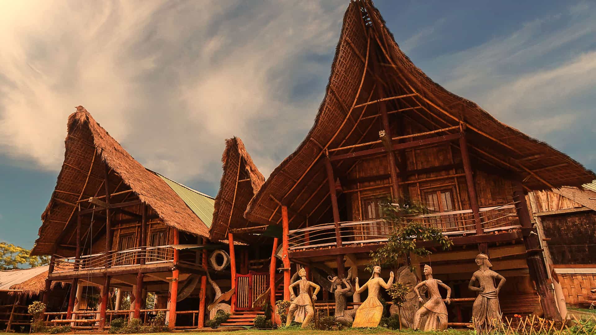 "A wide-angle, low-angle shot of traditional bamboo stilt houses with thatched roofs in a village on Majuli Island, India, under a dramatic cloudy sky.  "