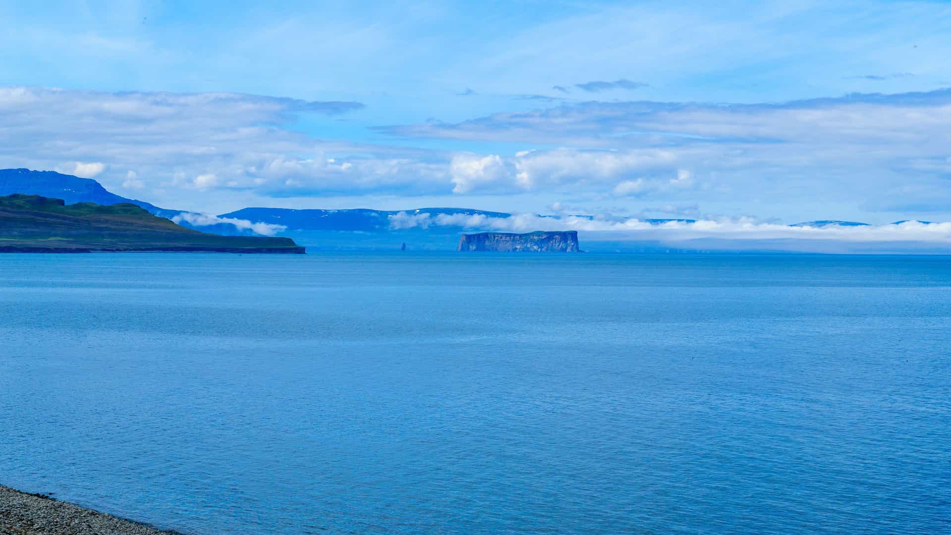A serene landscape view of Malmey Island, Iceland, with a flat-topped island rising from the sea, surrounded by misty mountains under a blue sky.