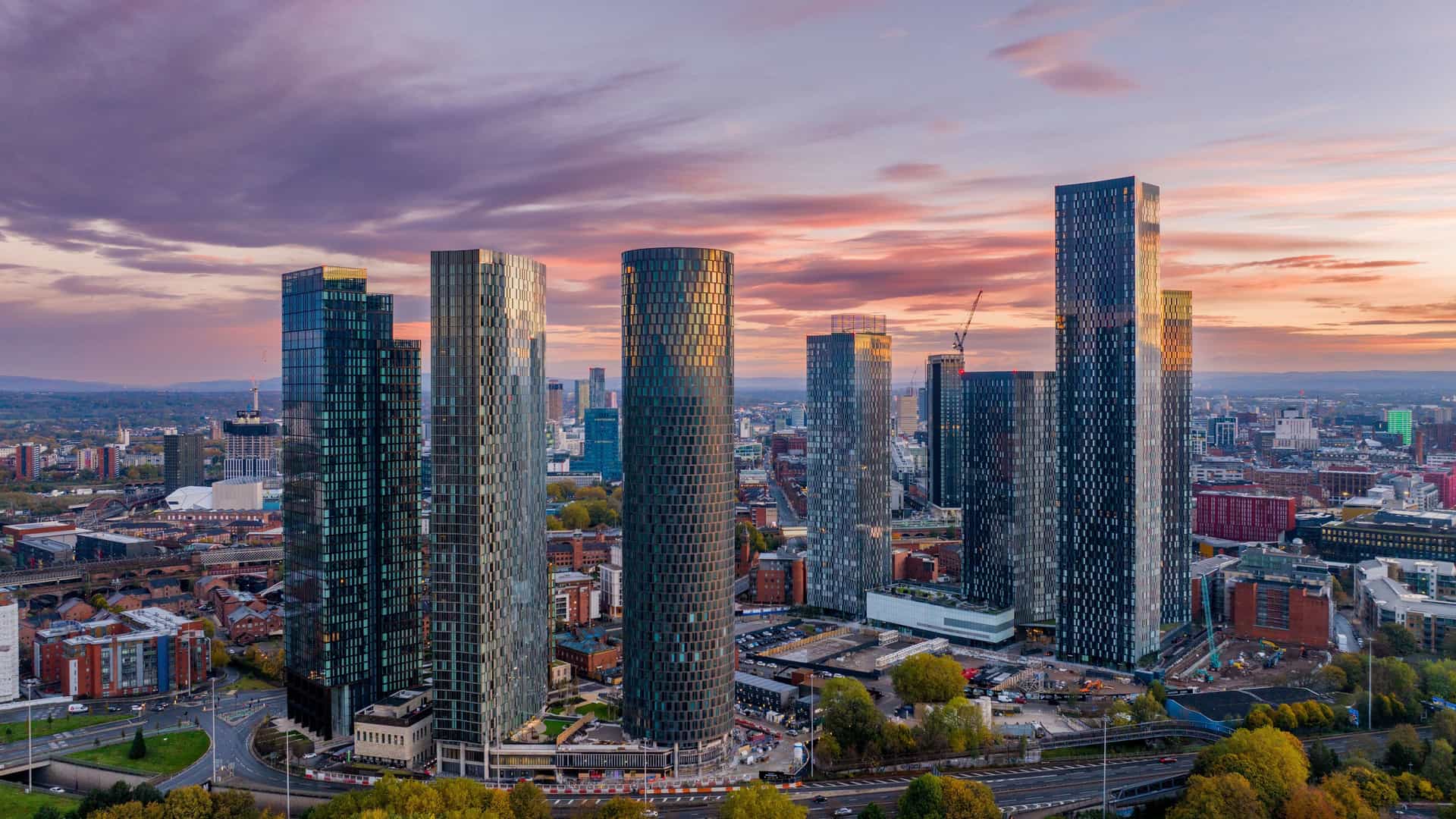 "An aerial sunset view of the modern residential skyscrapers of Deansgate Square in Manchester, England, reflecting the vibrant pink and orange clouds.  "