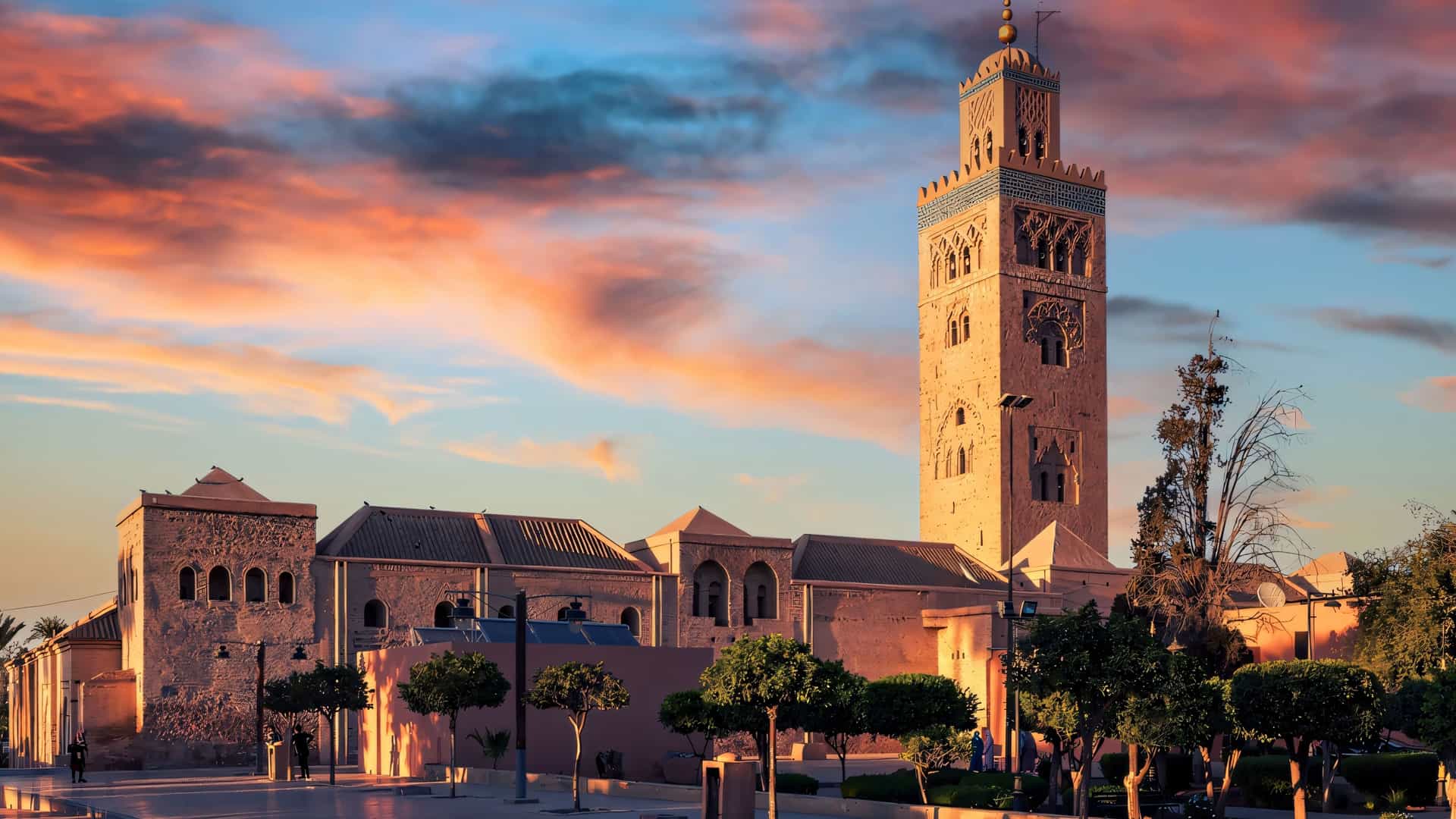 "A wide-angle sunset view of the Koutoubia Mosque in Marrakech, Morocco, with its prominent minaret silhouetted against a vibrant orange and blue sky.  "