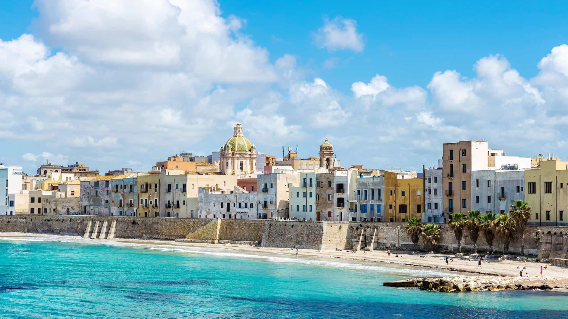 "A coastal view of the city of Marsala, Italy, with historic buildings and a prominent cathedral dome overlooking the turquoise Mediterranean Sea on a sunny day.  "