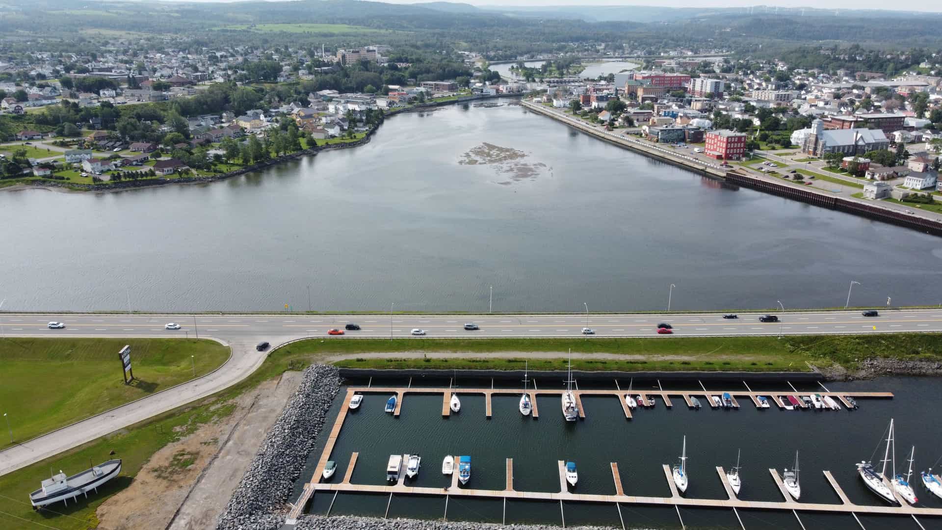 "An aerial view of the city of Matane, Canada, showing the river and marina with boats, a highway, and the town's buildings and rolling hills in the background.  "