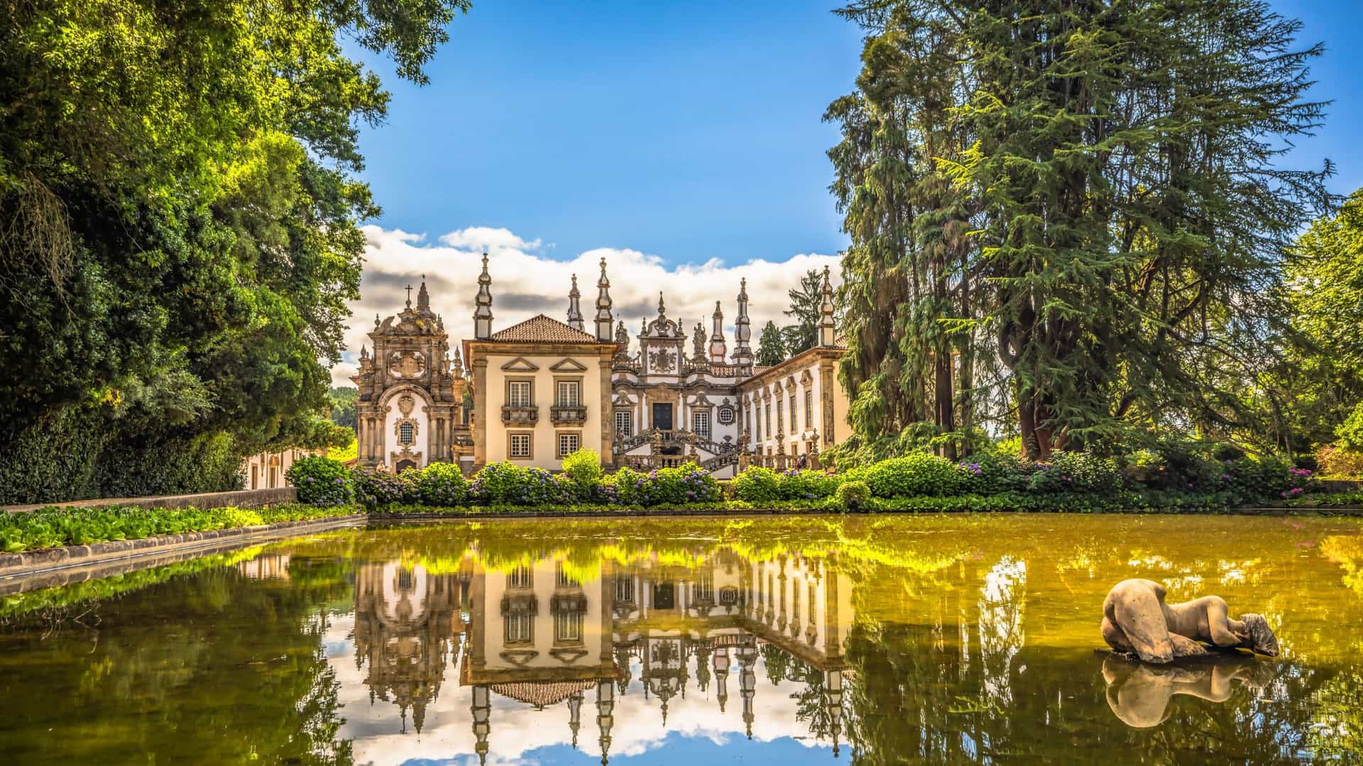 A picturesque view of the Baroque-style Casa de Mateus palace in Portugal, with its ornate facade and garden reflecting on a tranquil pond with a statue.