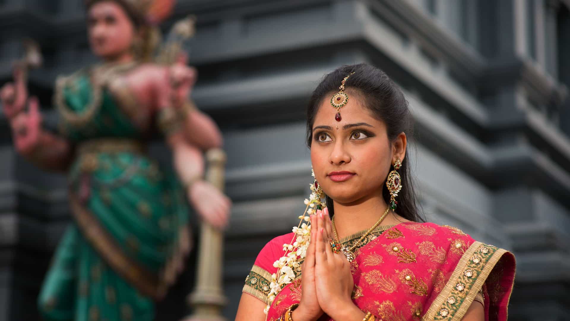 A portrait of a young Indian woman in a traditional pink sari with a bindi and jewelry, her hands clasped in a gesture of prayer, with a statue of a deity blurred in the background, a sight from Matiari.
