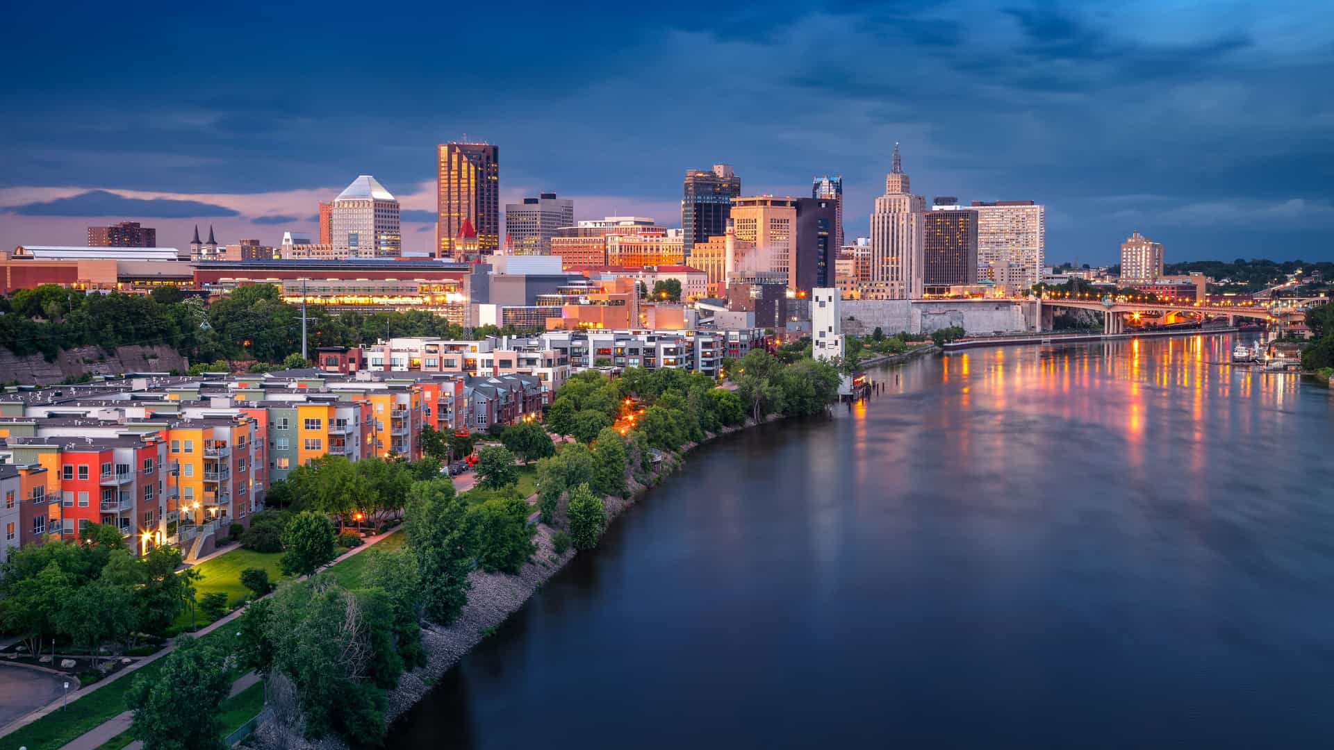 "An aerial night shot of the Minneapolis, Minnesota, skyline, with the Mississippi River reflecting the illuminated downtown buildings, colorful residential apartments, and bridges.  "