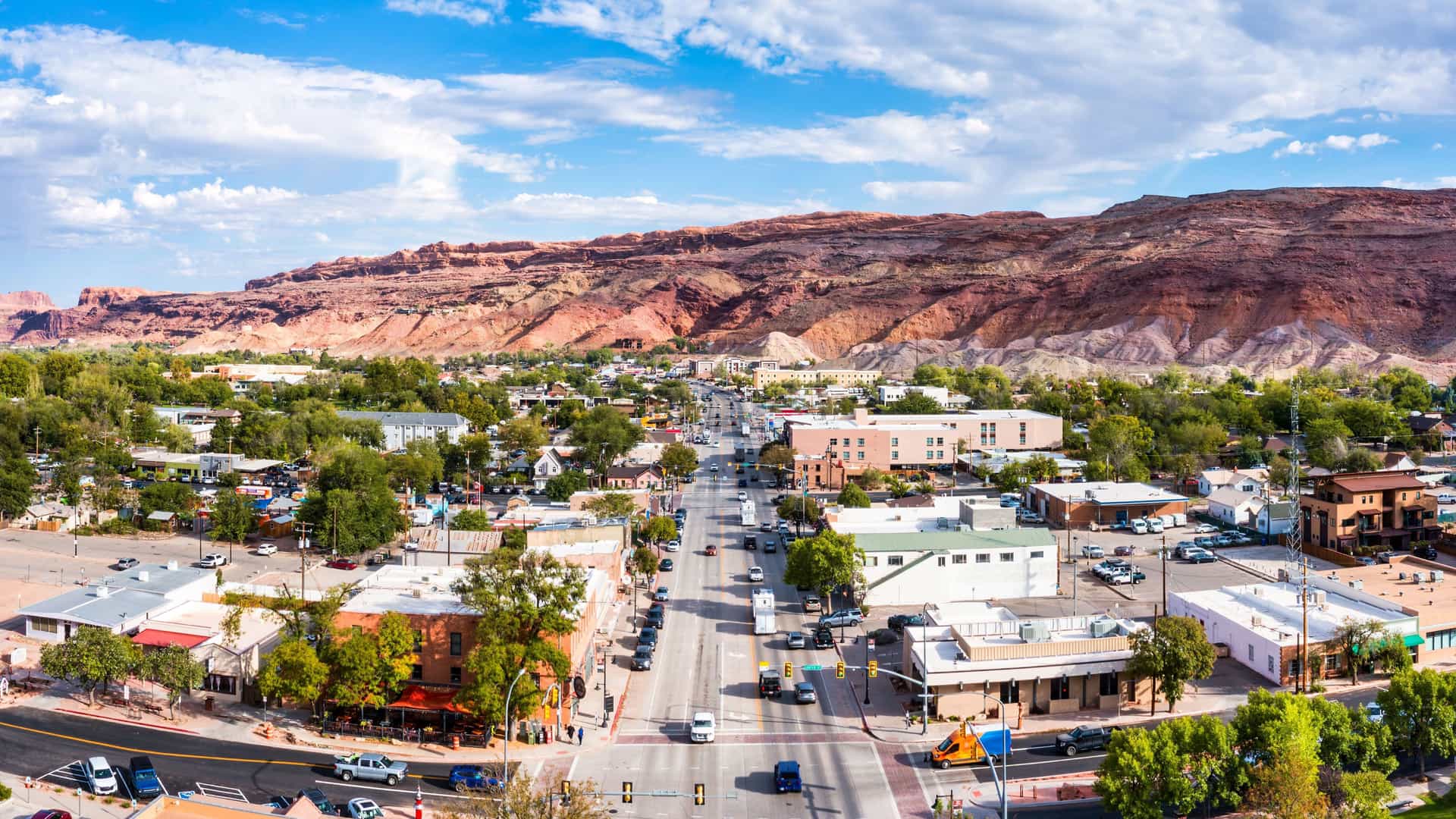 "An aerial panoramic view of downtown Moab, Utah, showing the main street, buildings, and surrounding landscape with the majestic red rock mountains in the background under a blue sky.  "