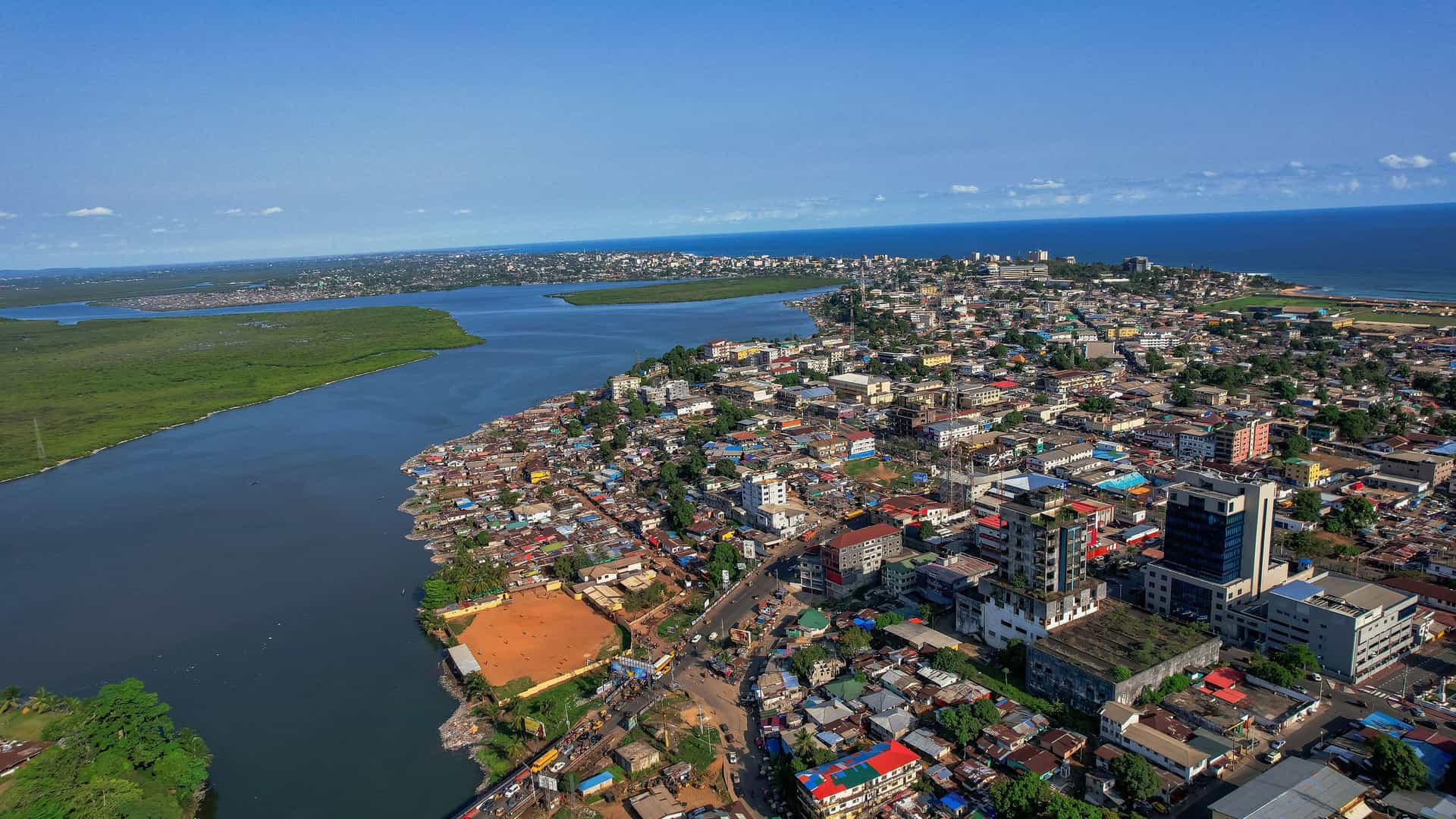 An aerial view of the coastal city of Monrovia, Liberia, showcasing a densely populated urban area, the Mesurado River, and the vast Atlantic Ocean meeting the shoreline.