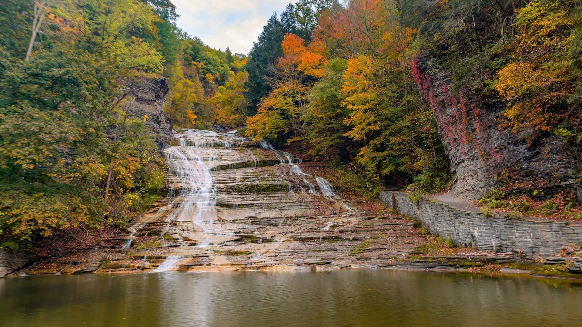 A wide, tiered waterfall cascades down rock ledges into a dark pool, surrounded by lush autumn foliage in red and gold.