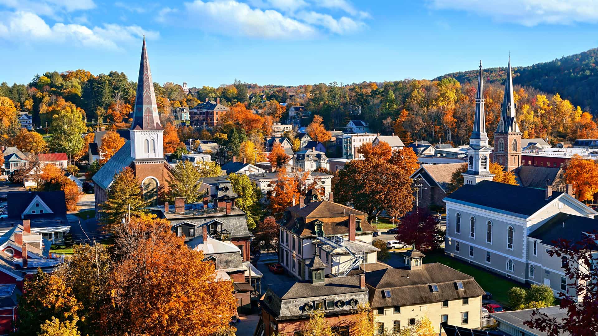 "An aerial autumn view of Montpelier, Vermont, with its historic buildings, two prominent church steeples, and vibrant fall foliage blanketing the surrounding hillsides.  "