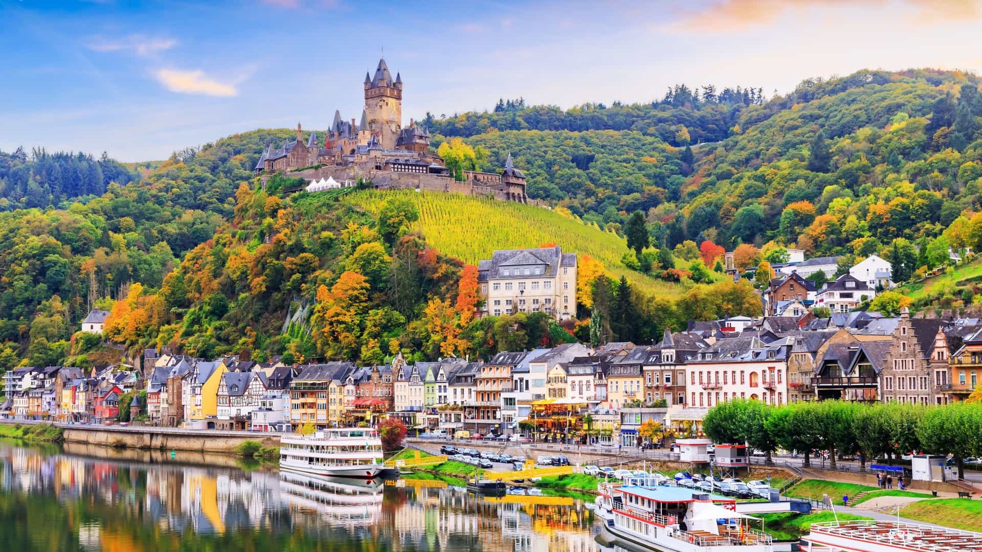 A picturesque view of Cochem, Germany, with the historic Reichsburg Castle perched on a hill above the colorful town and the Moselle River, with cruise ships docked along the embankment.